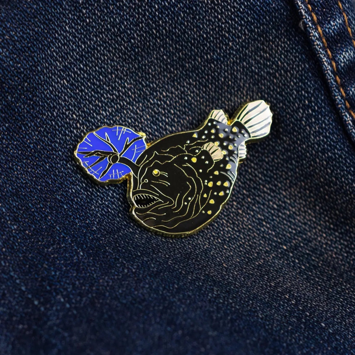 Stemcell Atlantic Football Fish enamel pin with blue lure displayed on dark blue denim. Unique ocean-themed collectible pin.