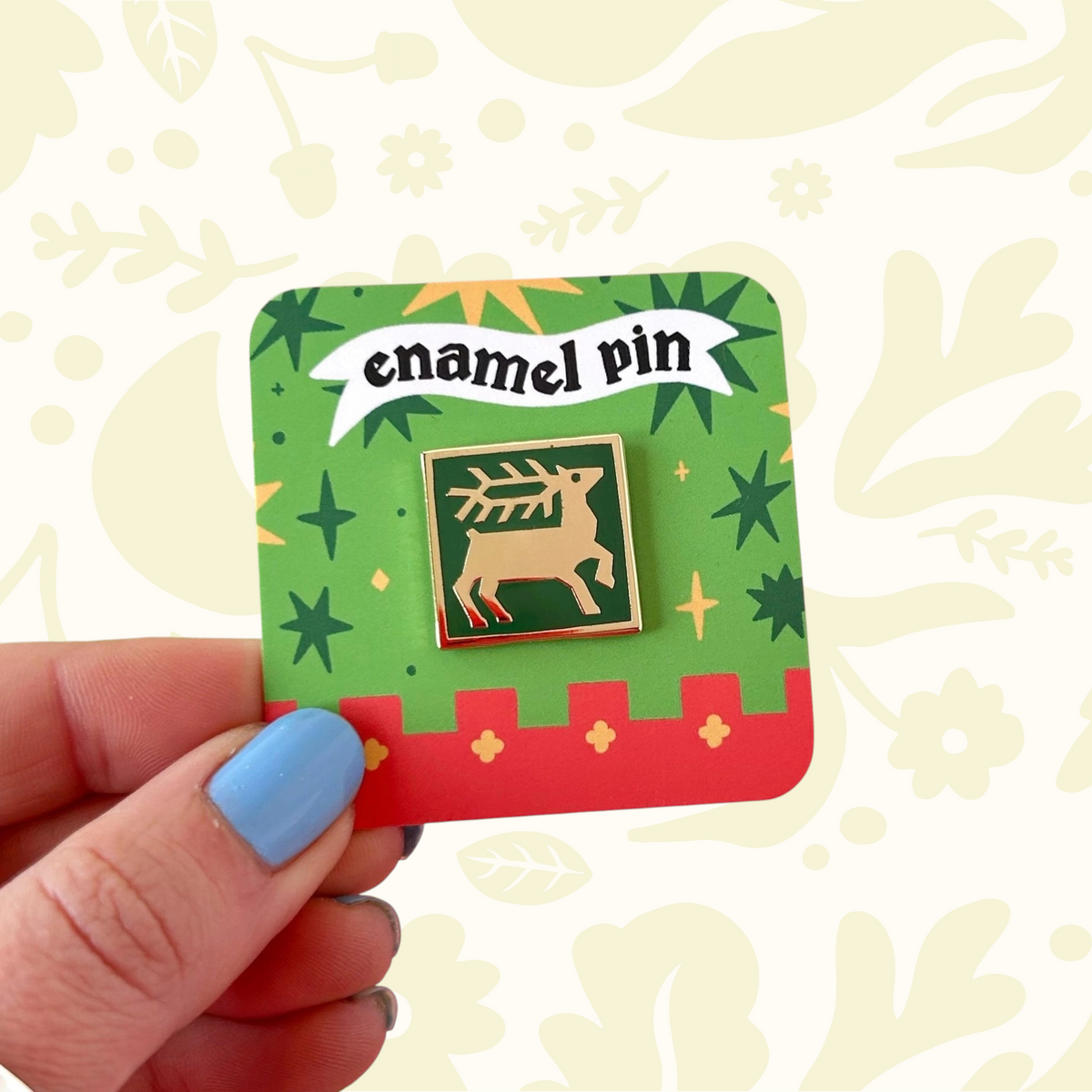 Blue-nailed hand displays Stacey McEvoy Caunt Deer Tile enamel pin (Aug 2026) on a vibrant green background.