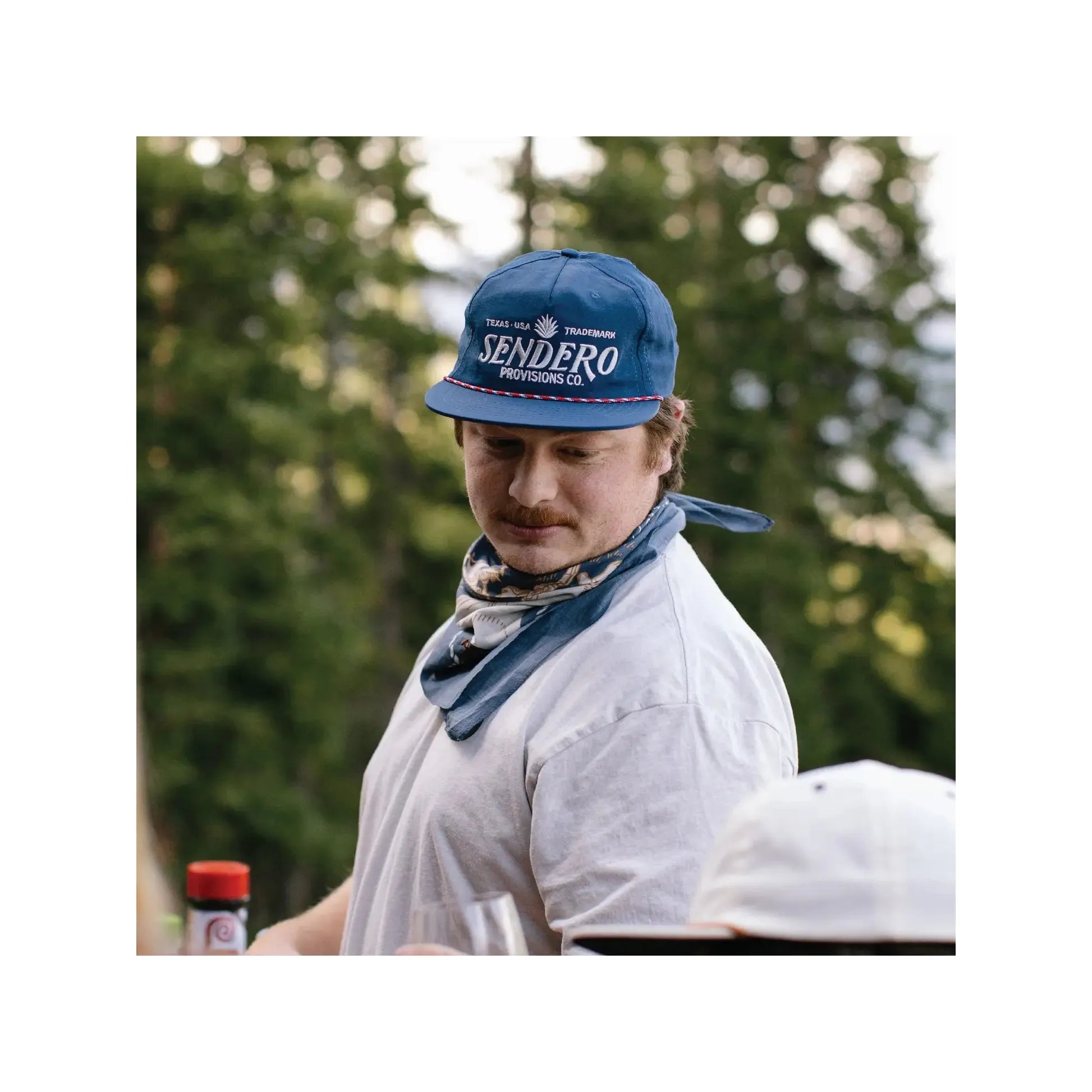 The Sendero Provisions Skeleton Riders Bandana is a navy blue accessory adorned with cowboy and Western-themed illustrations, featuring the text "Sendero Provisions" in the center. It is crafted from 100% Organic Japanese Cotton.