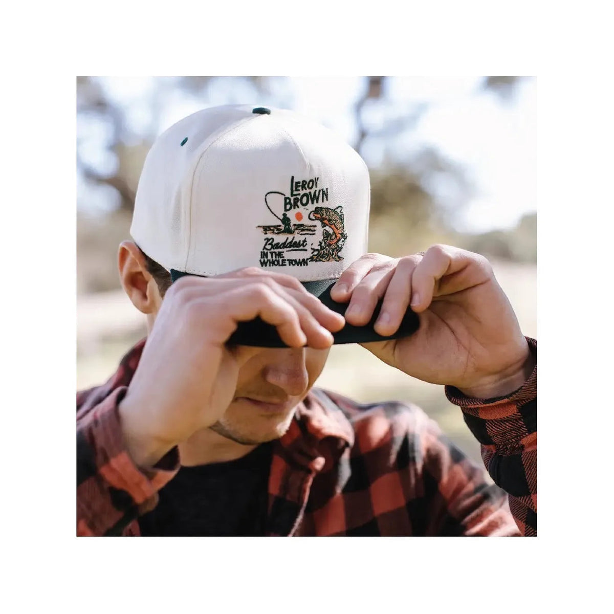 A person outdoors adjusts the brim of their Sendero Provisions LeRoy Brown Cap, wearing a red plaid shirt.