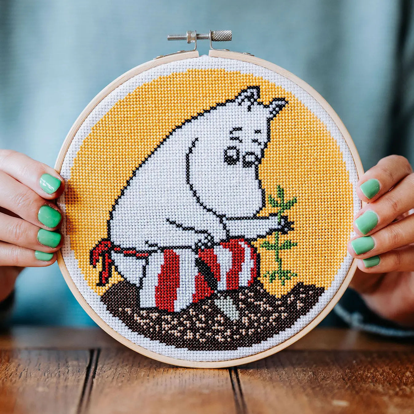A person with green nail polish holds The Crafty Kit Company Moominmamma Gardening Cross Stitch, framed in a hoop, depicting Moominmamma sitting on a striped pillow in a garden.