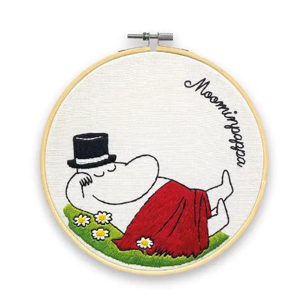 The Crafty Kit Company Moominpapa Snoozing Cross Stitch features an embroidery of Moominpappa resting on grass with daisies, covered by a red blanket, in a wooden hoop with "Moominpappa" written above. This charming cross stitch kit includes all the necessary embroidery supplies to recreate this cozy scene.