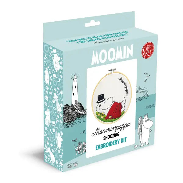 The Crafty Kit Company Moominpapa Snoozing Cross Stitch features an embroidery of Moominpappa resting on grass with daisies, covered by a red blanket, in a wooden hoop with "Moominpappa" written above. This charming cross stitch kit includes all the necessary embroidery supplies to recreate this cozy scene.