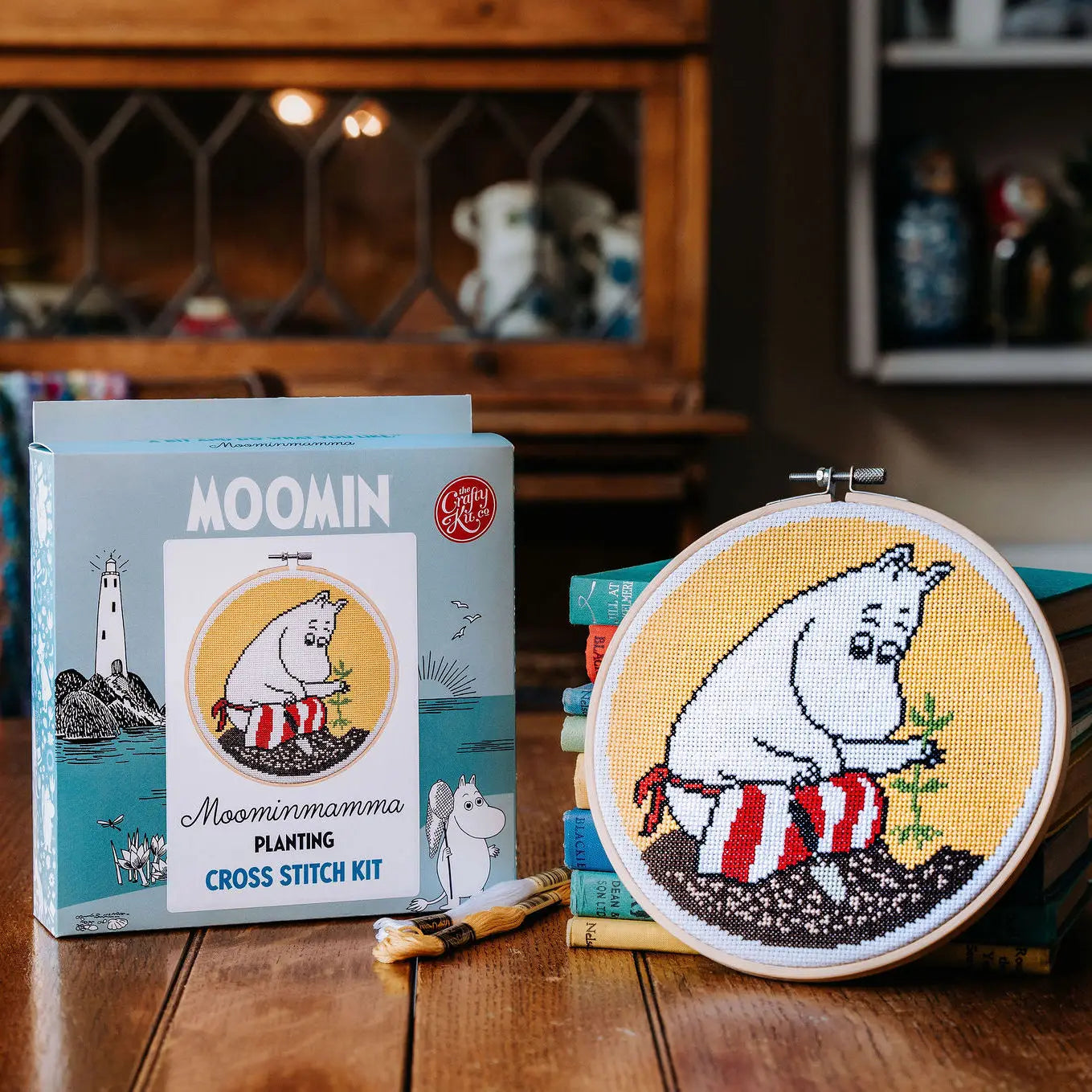 A The Crafty Kit Company Moominmamma Gardening Cross Stitch kit is showcased on a wooden table, featuring vibrant embroidery thread and the finished piece tastefully framed in a hoop next to its charming packaging.
