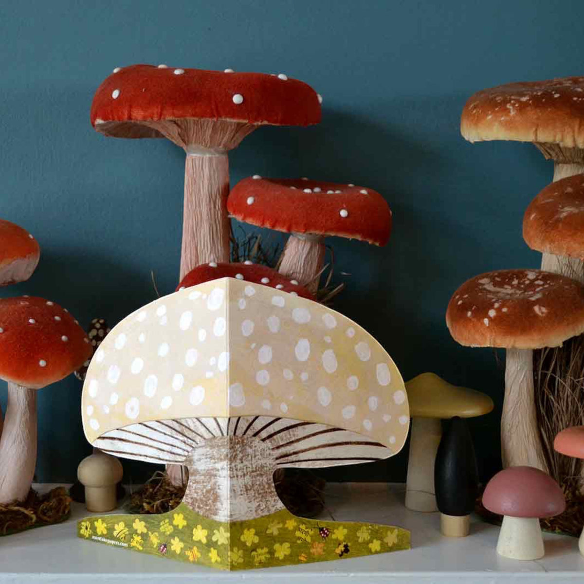 Mushroom shaped greeting card surrounded by an assortment of mushroom decor on a white shelf.