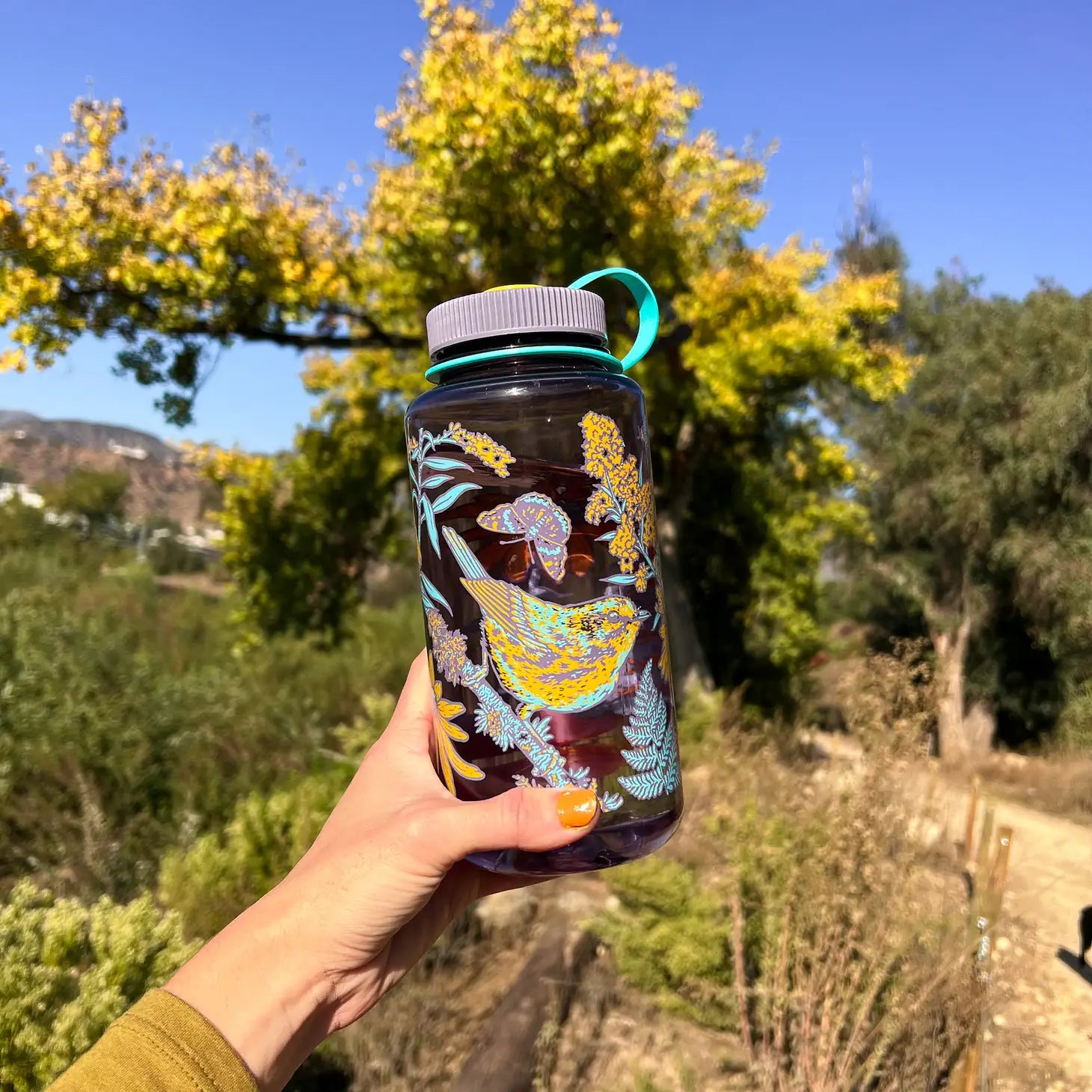 Introducing the limited edition Mustard Beetle Prairie 32oz Wide Mouth Nalgene Water Bottle, featuring a charming design adorned with birds, flowers, and bees. This eco-conscious water bottle is crafted from BPA-free recycled plastic and comes displayed on a wooden surface with an emblem tag attached.
