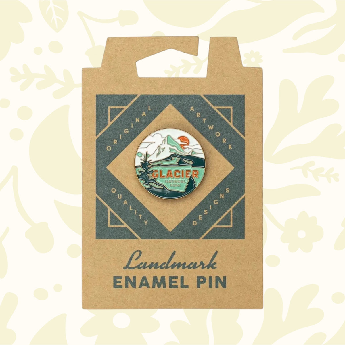 A round enamel pin featuring a glacier scene with mountains and text saying "GLACIER NATIONAL PARK" is attached to a brown cardboard backing made from sustainable packaging, sold under the name The Landmark Project Glacier National Park Pin.