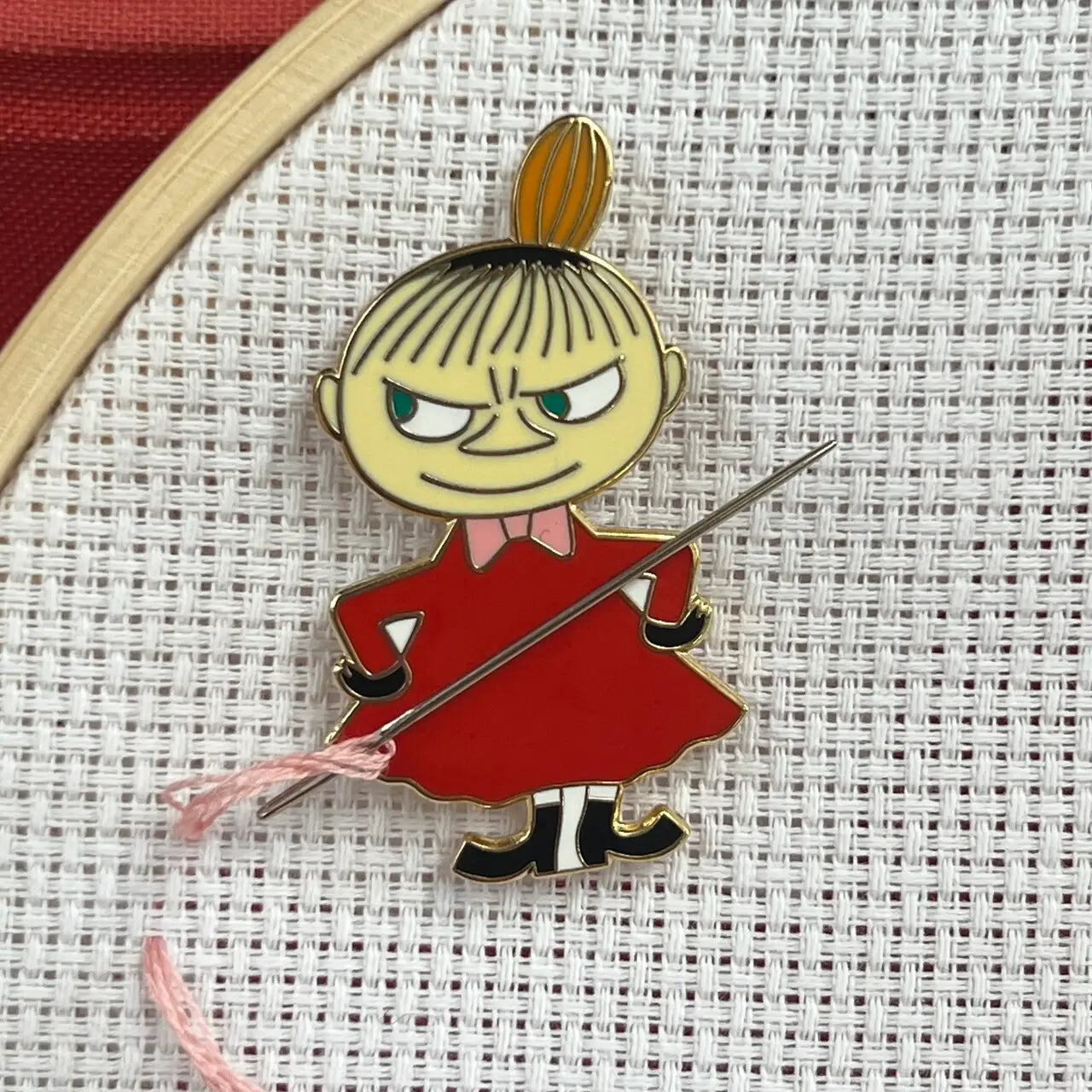 The Crafty Kit Co Moomin Needle Minder features a Little My character with a mischievous expression, dressed in red and holding a needle with pink thread. This charming embroidery accessory comes equipped with a strong magnet to keep your project secure.