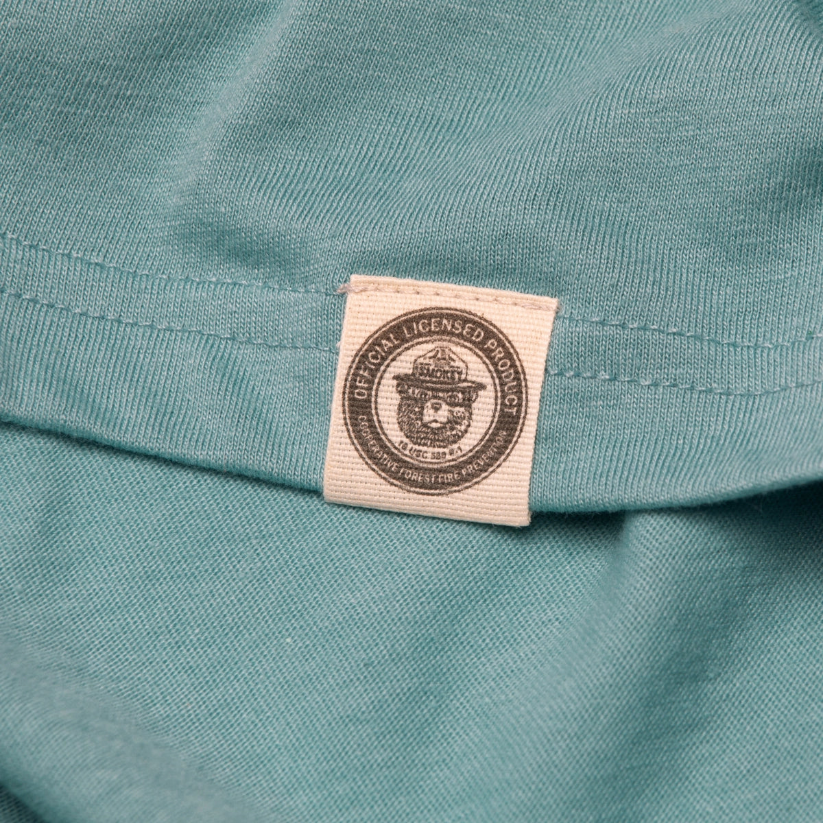 Close-up of a small label on a green fabric. The label, attached to *The Landmark Project Ringer Smokey Bear T-Shirt - Glacier*, reads "OFFICIAL LICENSED PRODUCT" with an emblem in the center.