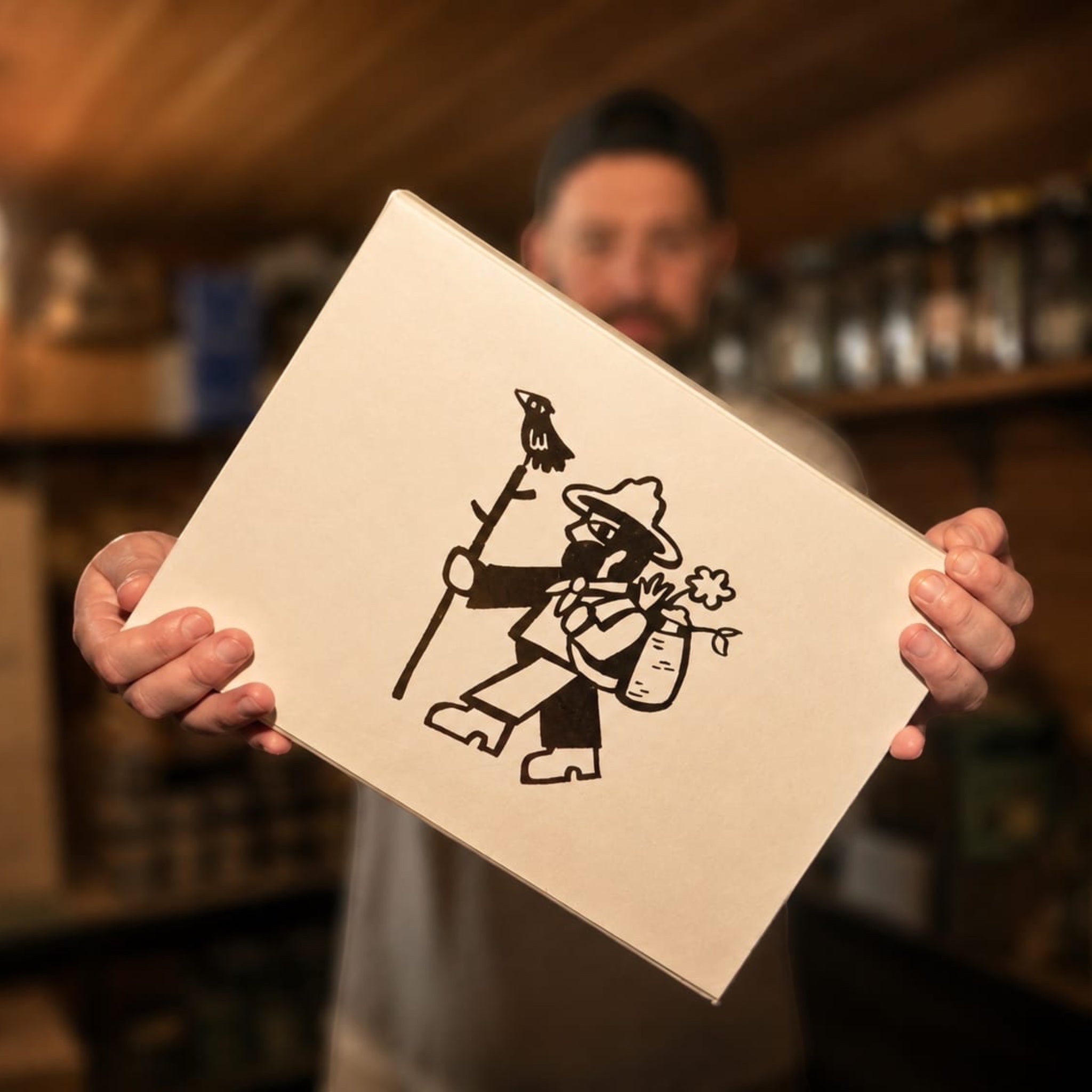 Person holding white box with hiker illustration—stick, sack, rustic outdoors; hiking gear gift packaging, adventure theme.