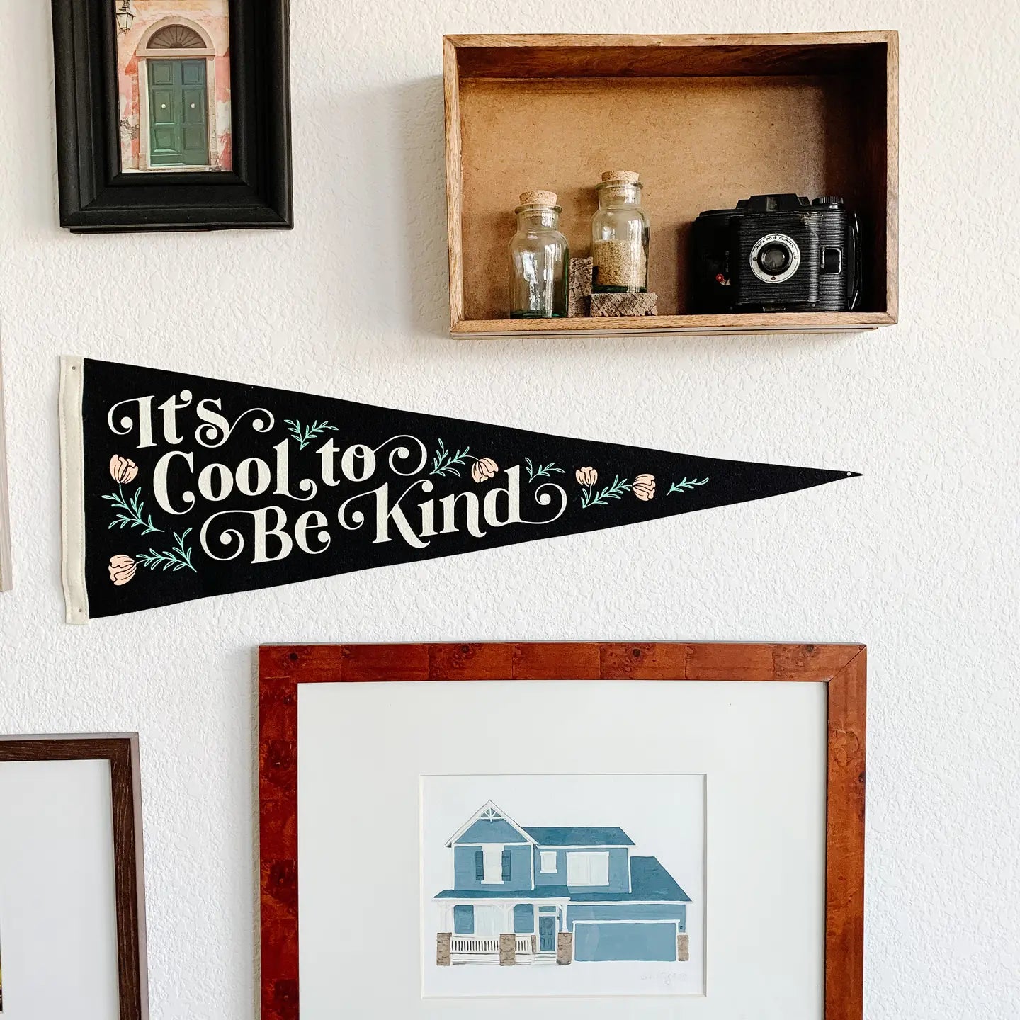 Gallery wall with framed art, glass jars, vintage camera, and It’s Cool To Be Kind Oxford Pennant home decor display.