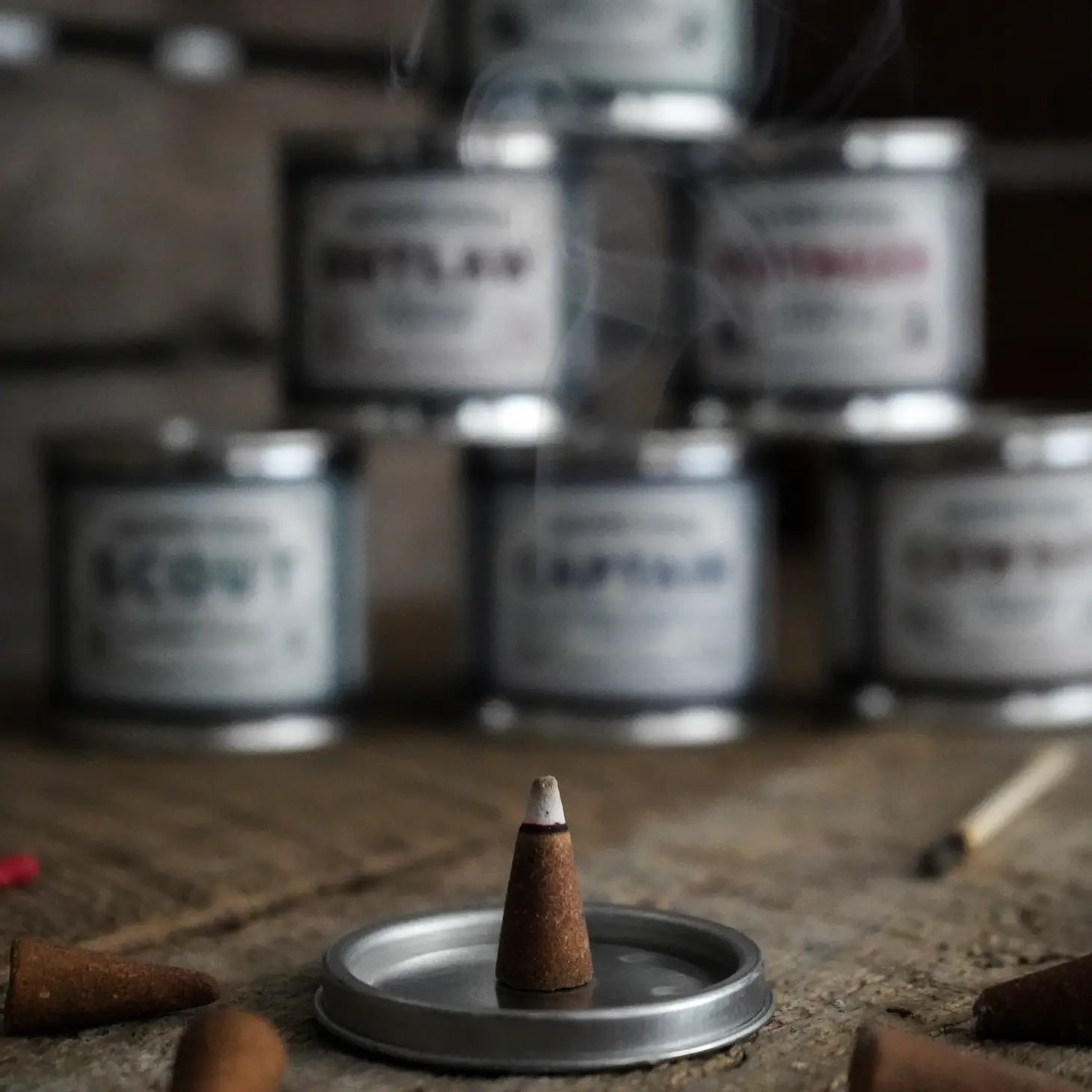 A burning Ambush Candle Co Incense Cone in Cinnamon & Vetiver - Outlaw rests on a metal lid, with blurred tin containers in the background. Smoke is gently rising from the cone, showcasing its American-made quality and 20+ minute burn-time.