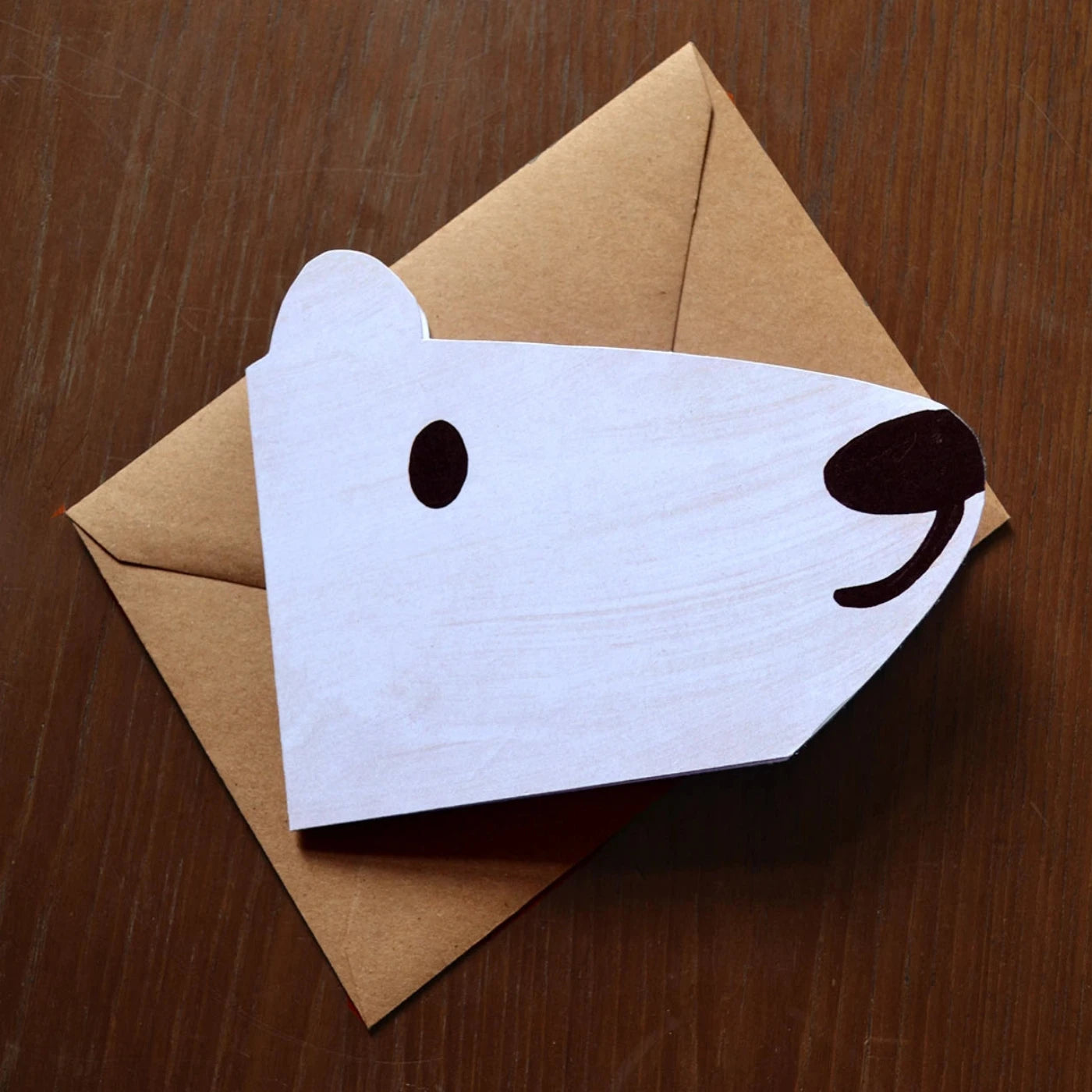 Face of a polar bear shaped card, laid on a brown envelope on a brown wooden table top.