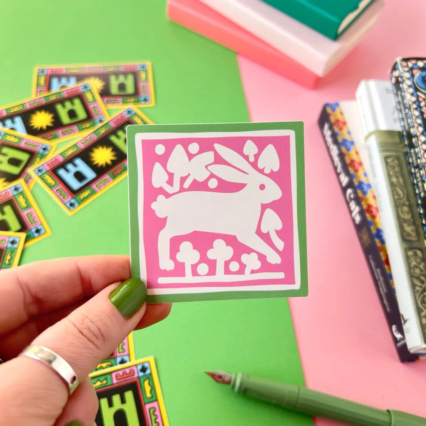 A hand displays a Medieval Rabbit Tile Sticker by Stacey McEvoy Caunt, featuring mushrooms, beside books and pens on a desk.