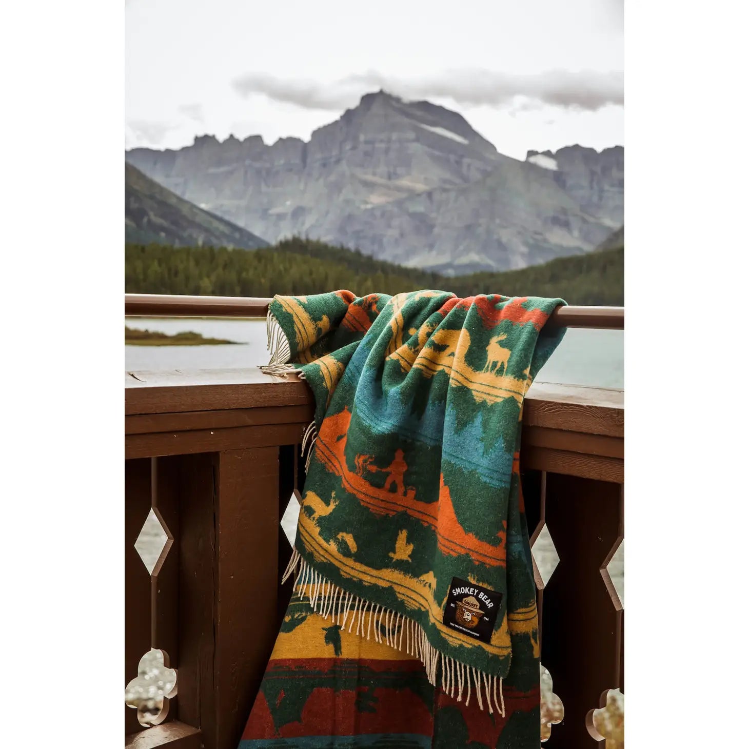 Introducing the Sackcloth & Ashes Smokey Bear Blanket - Smokey Wild: a uniquely crafted blanket featuring a vibrant forest and wilderness pattern adorned with mountains, wildlife, and trees in blue, red, and yellow hues. This special piece promotes wildfire prevention while being thoughtfully made from recycled materials.