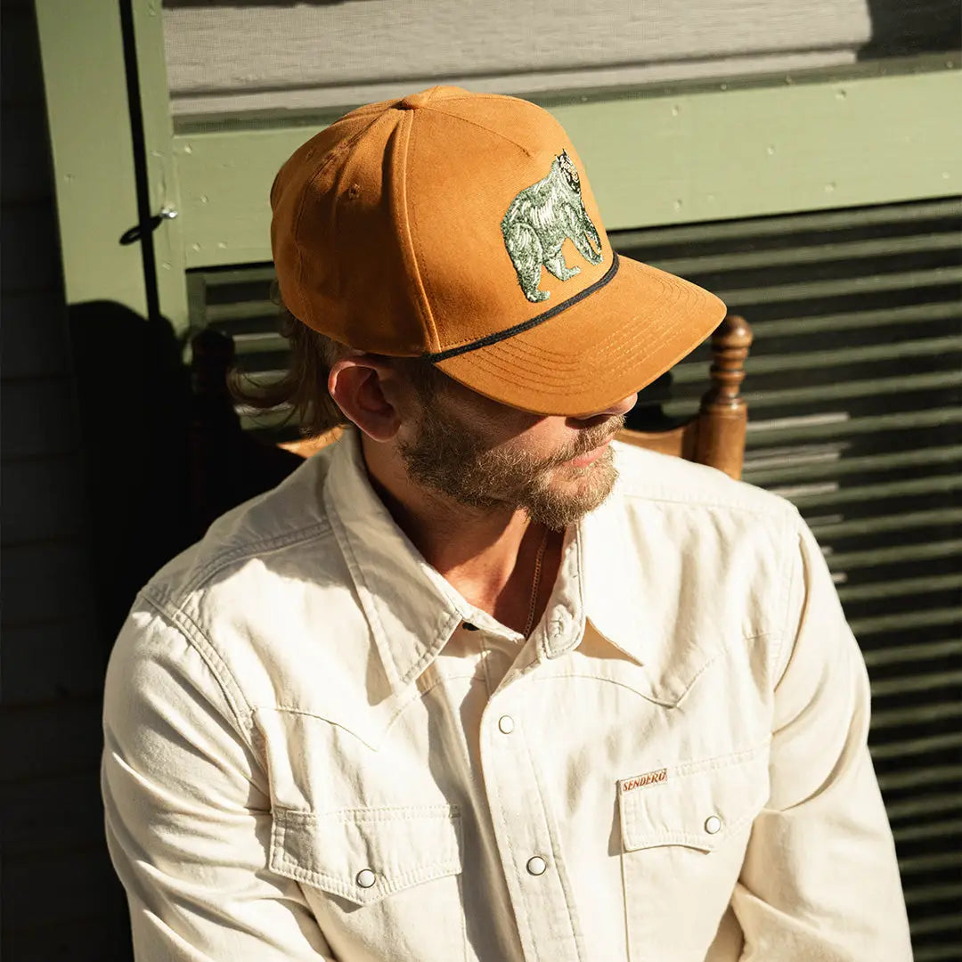 Man wearing a Sendero Bear Graphic Cap and cream button-up sits sideways on a wooden chair, relaxed pose for casual style.
