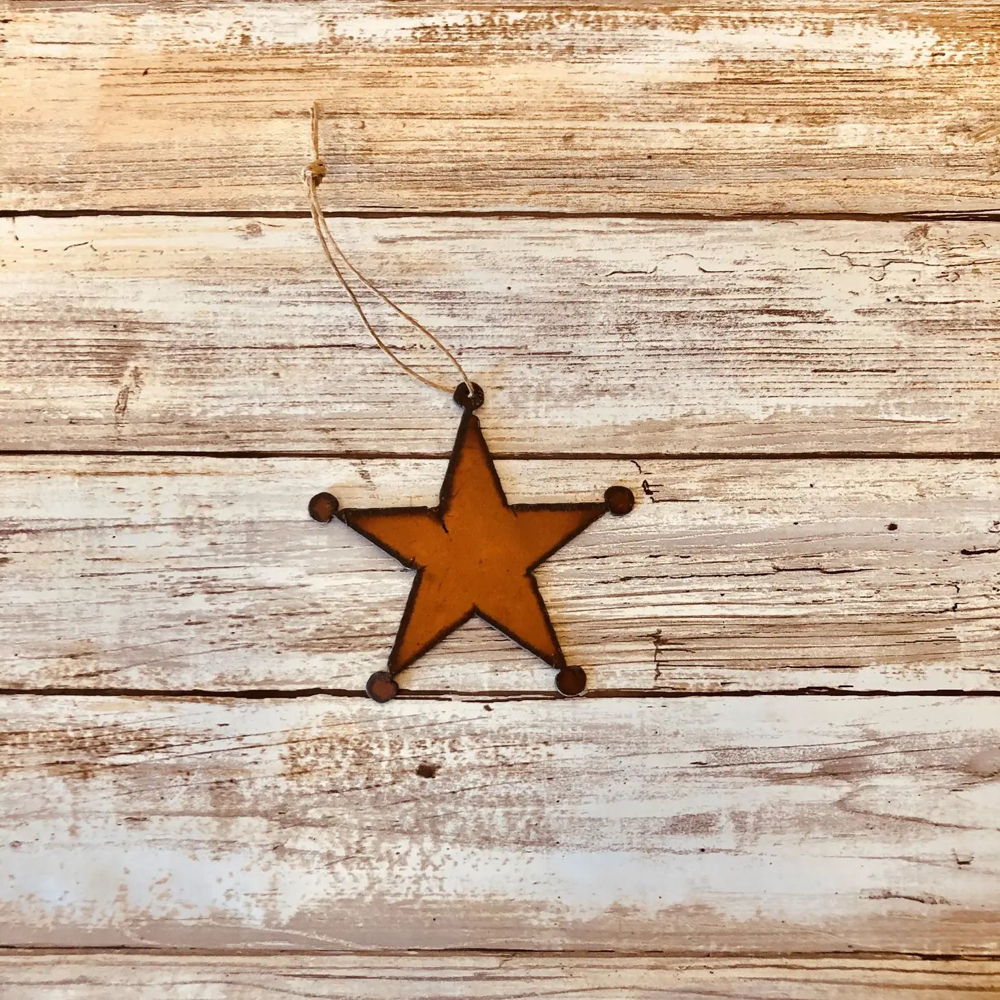 A Universal Iron Works Sheriff Star Hanging Decoration, expertly crafted from 18 gauge steel, dangles by a string against a weathered wooden background, highlighting its natural rusting process.