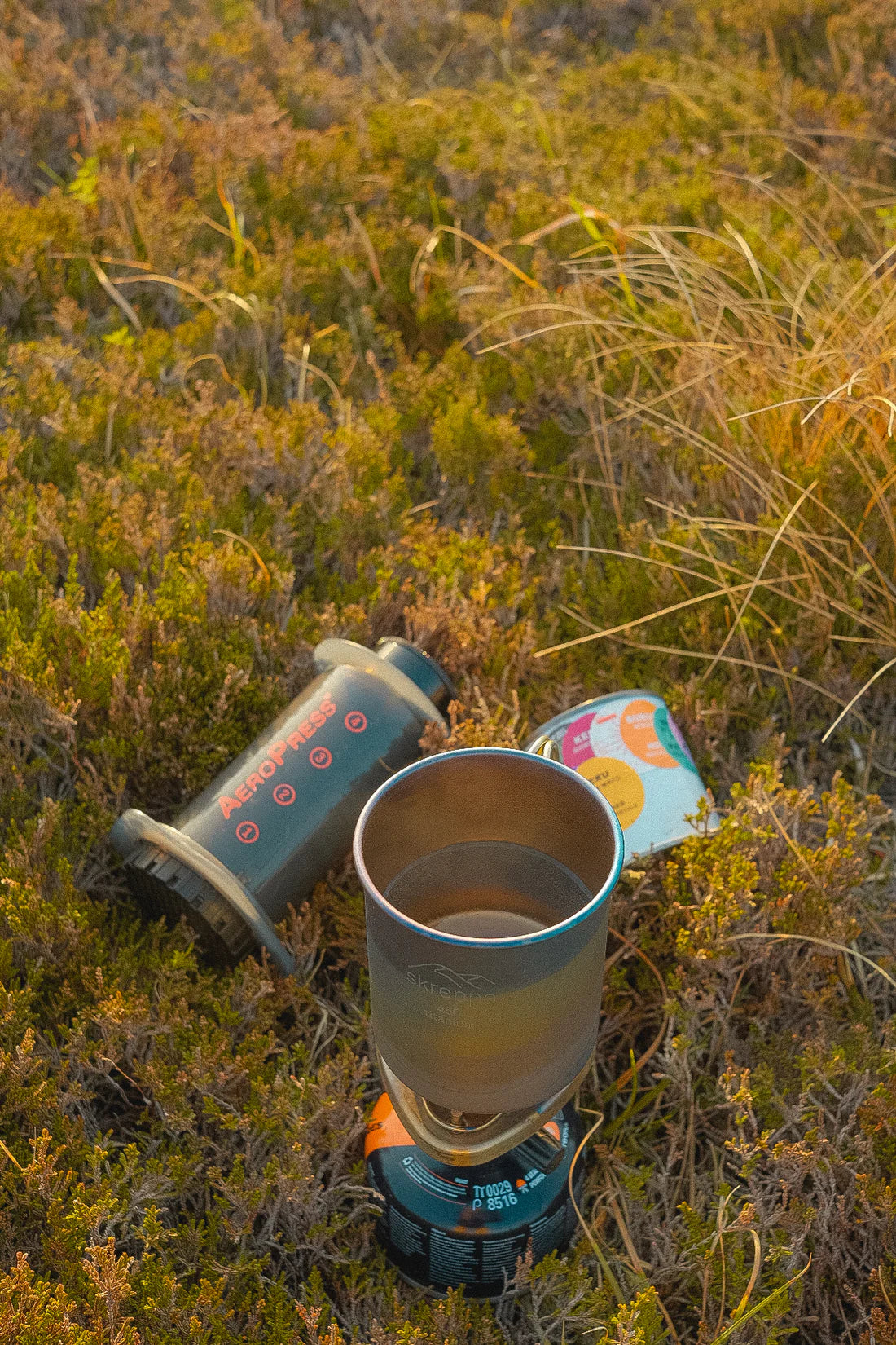 Ultralight Skreppa Single Wall Titanium Mug 450ml with AeroPress and food can on dry grass—perfect for outdoor coffee camping.