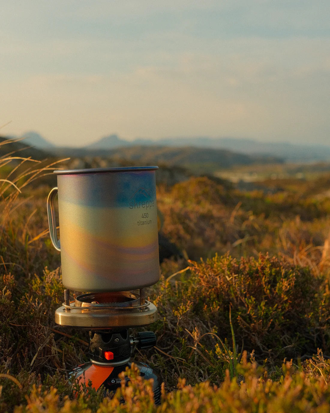 Skreppa Single Wall Titanium Mug 450ml Geodic on camp stove, grassy hills, scenic mountains—outdoor adventure camping gear.