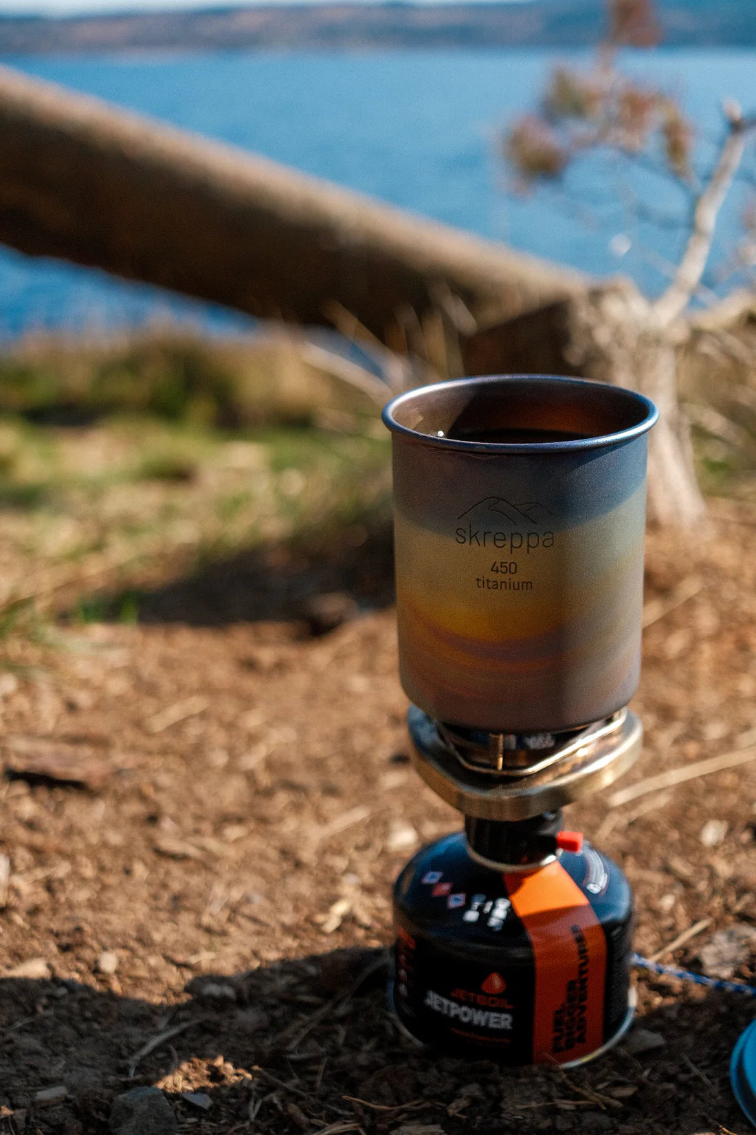 The Skreppa 450ml Titanium Mug-Geodic heats on portable stove near water, surrounded by trees; ideal for outdoor camping.