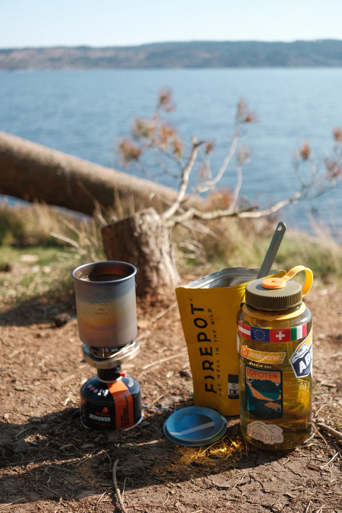 Skreppa Single Wall Titanium Mug 450ml Geodic beside lake, outdoor camping gear with scenic hills in background.