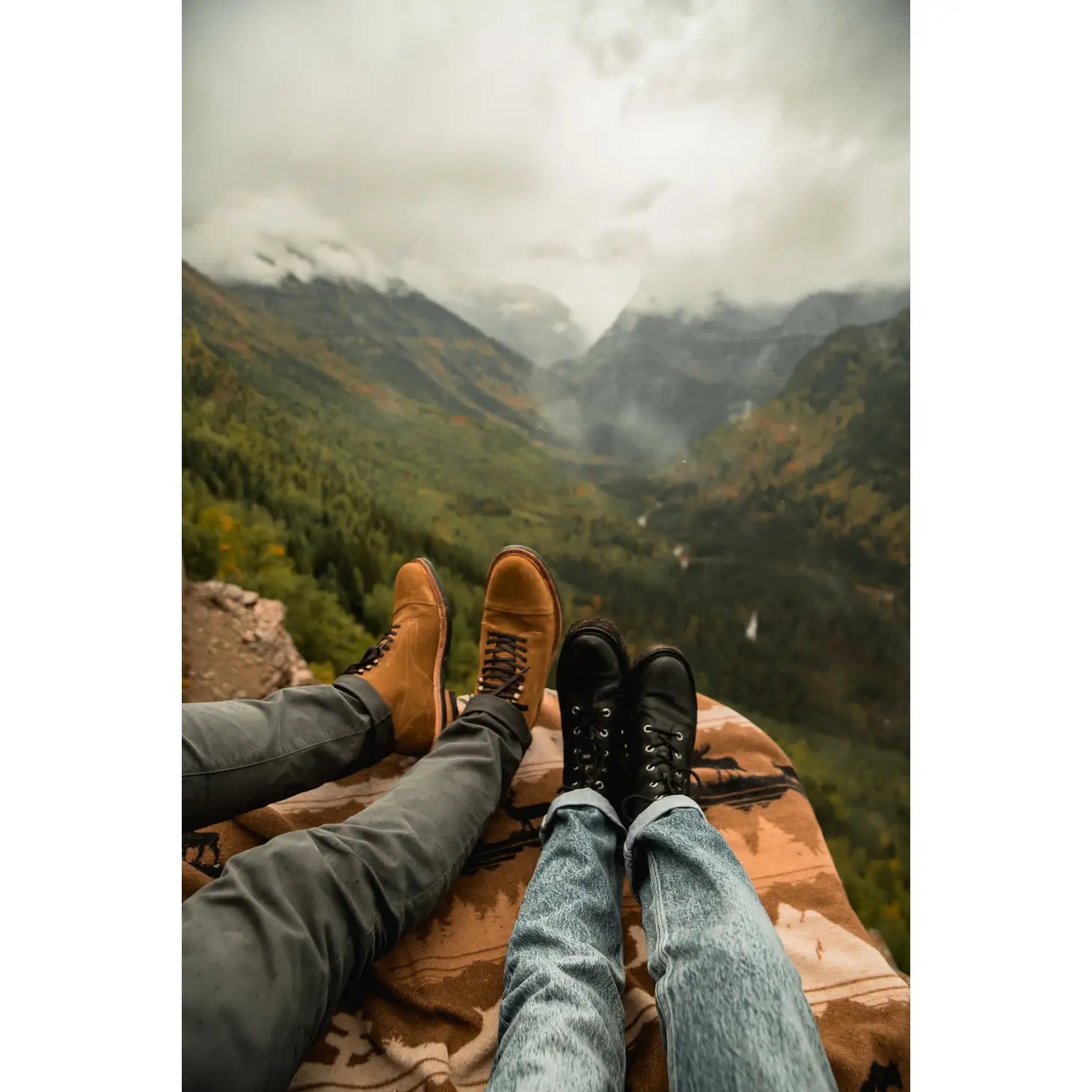 Two people relaxing on a Sackcloth & Ashes Smokey Bear Blanket - Smokey Roam with their legs stretched out, overlooking a scenic mountain valley shrouded in mist. With Smokey Bear's message of wildfire prevention in mind, they enjoy the serene beauty responsibly.