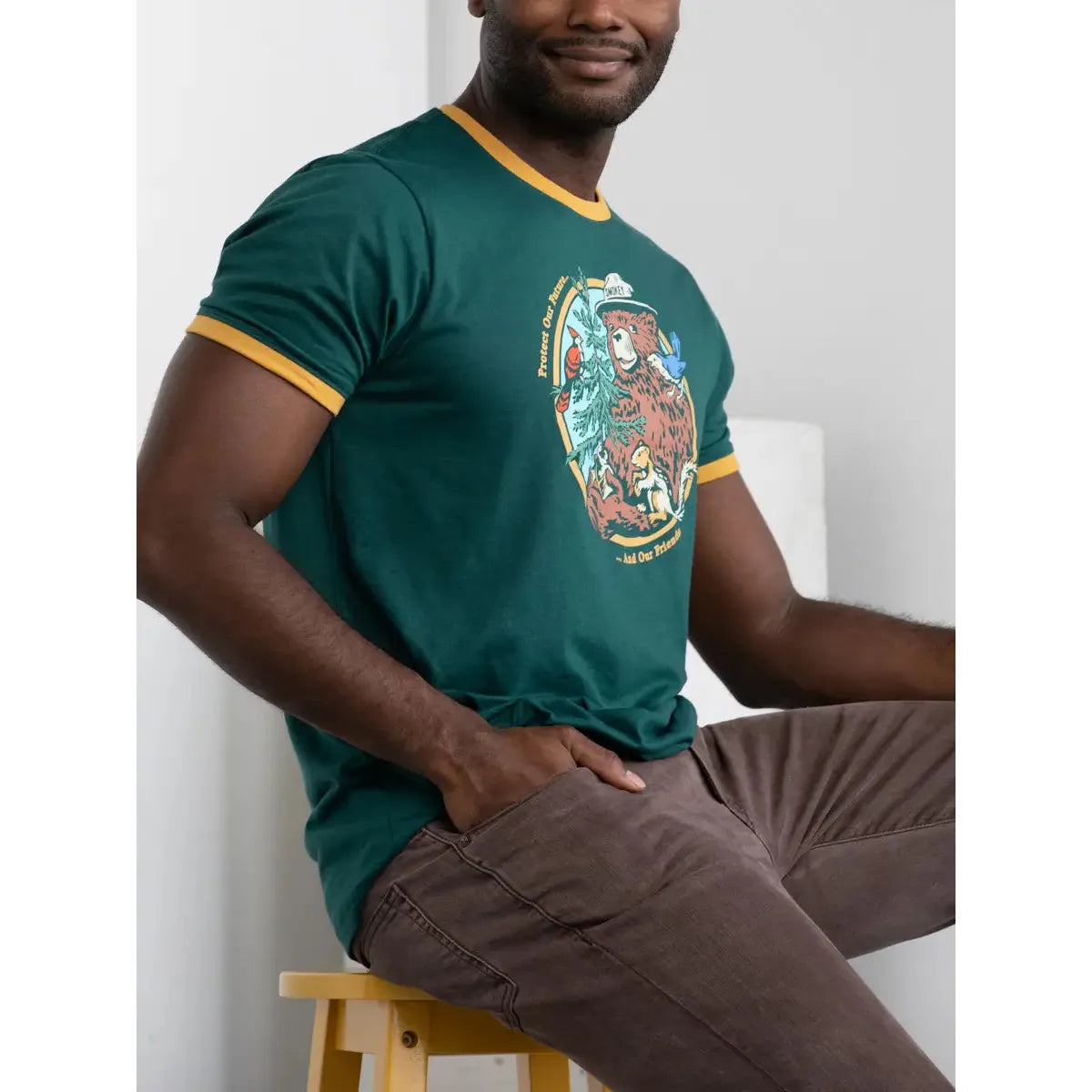 The Landmark Project Ringer Smokey Bear T-Shirt in Spruce showcases a green retro style with yellow trim, featuring an illustration of Smokey Bear wearing a hat, surrounded by trees and animals, and accompanied by text.