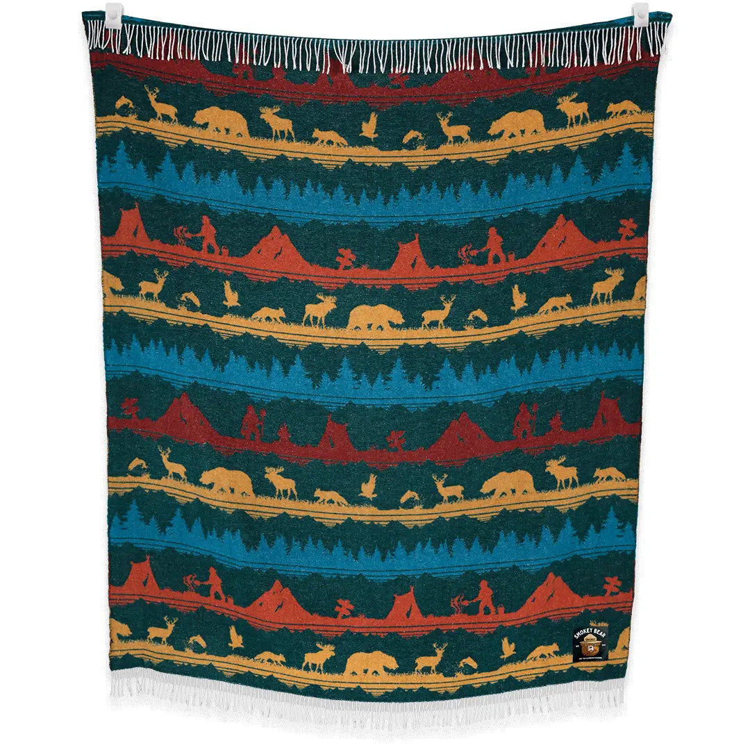 Introducing the Sackcloth & Ashes Smokey Bear Blanket - Smokey Wild: a uniquely crafted blanket featuring a vibrant forest and wilderness pattern adorned with mountains, wildlife, and trees in blue, red, and yellow hues. This special piece promotes wildfire prevention while being thoughtfully made from recycled materials.