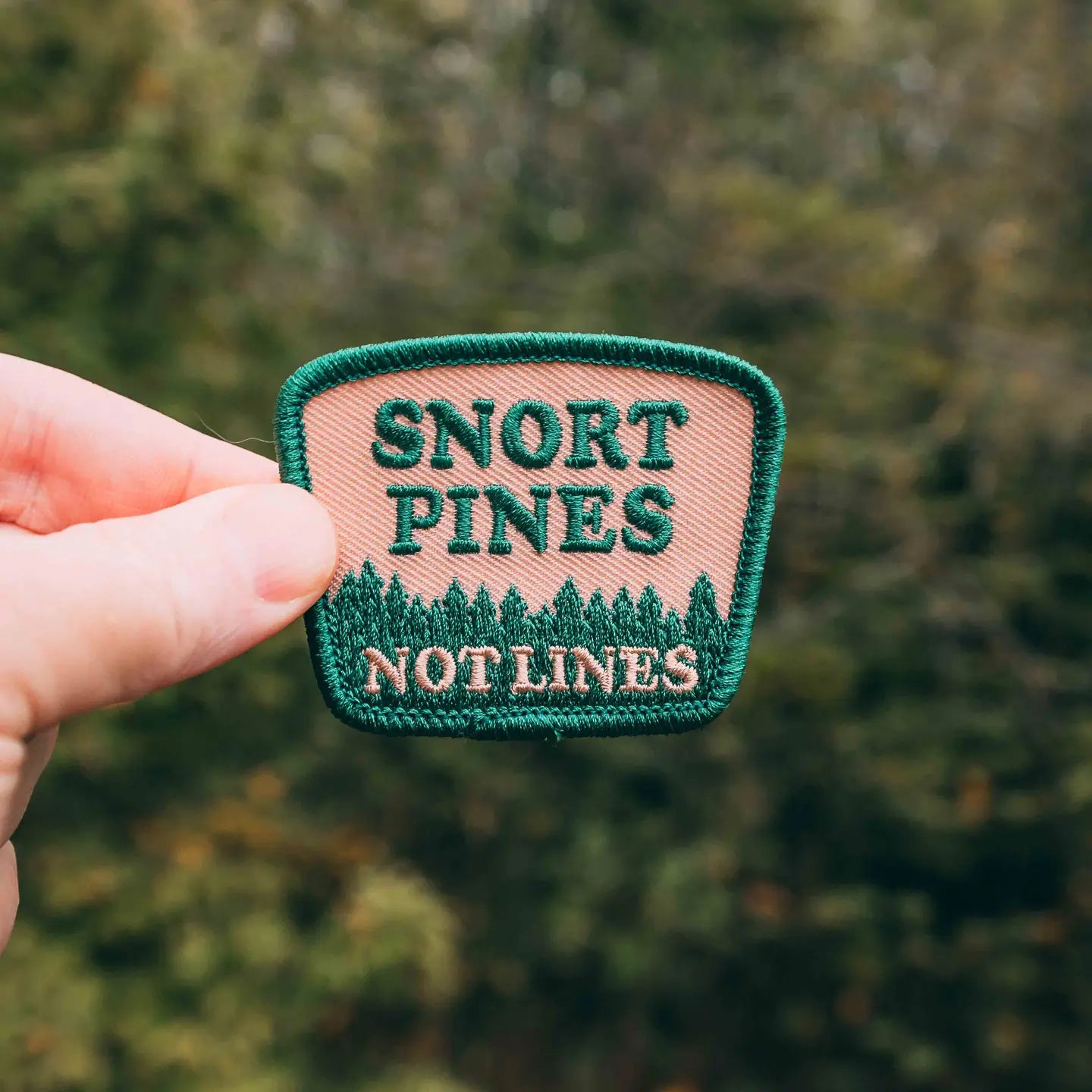 Hand holding Hey Bear Outdoors Snort Pines Not Lines embroidered hiker patch, pine tree design, outdoor hiking gear accessory.