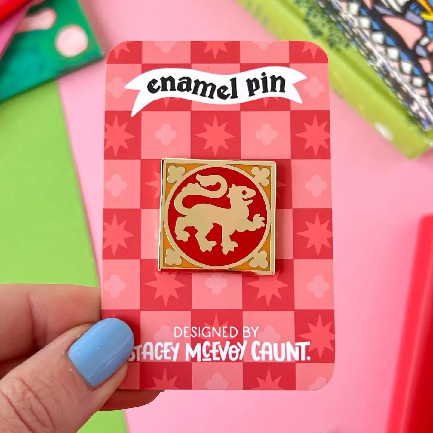 Hand displays Stacey McEvoy Caunt Lion Medieval Tile Pin, gold lion on red enamel, with red checkered backing card.