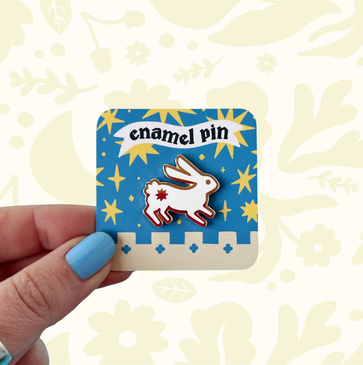 Hand holds Stacey McEvoy Caunt Medieval Rabbit Enamel Pin with blue and yellow star background, collectible accessory.