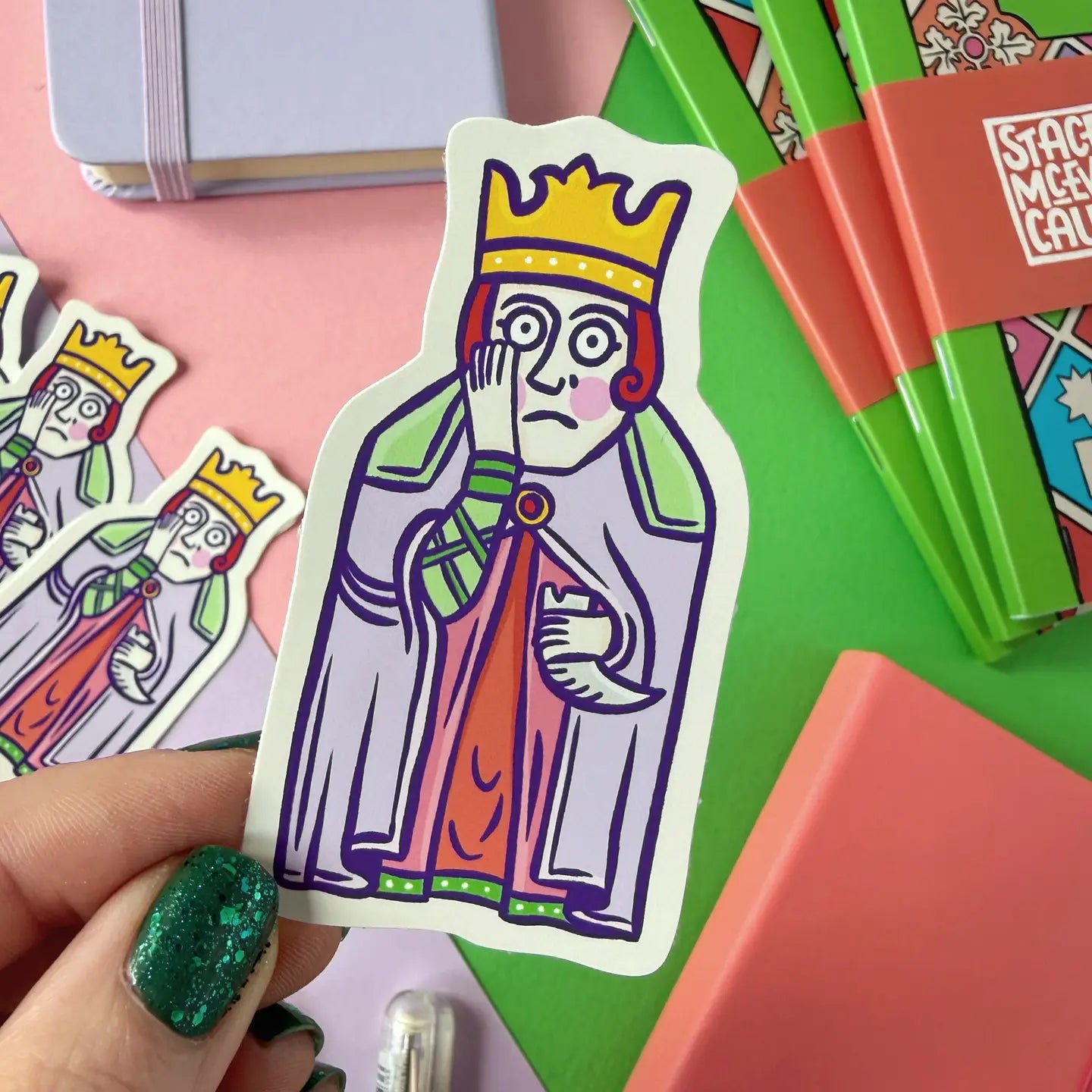 Hand displays a Stacey McEvoy Caunt Medieval Queen Lewis Chessmen vinyl sticker beside notebooks and collectible stickers.