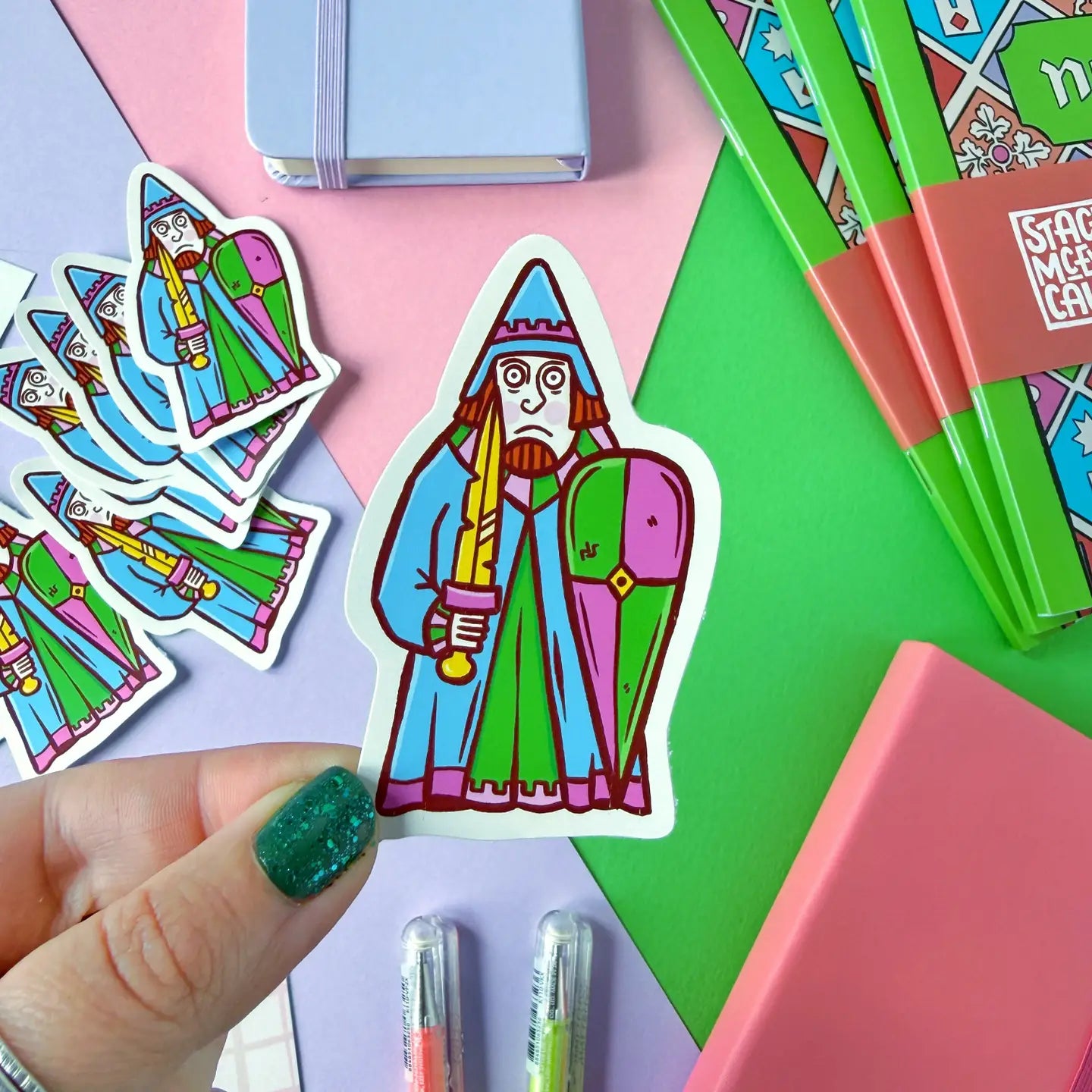 Hand holding a Stacey McEvoy Caunt Medieval Rook Lewis Chessmen sticker with colorful pens and notebooks in the background.
