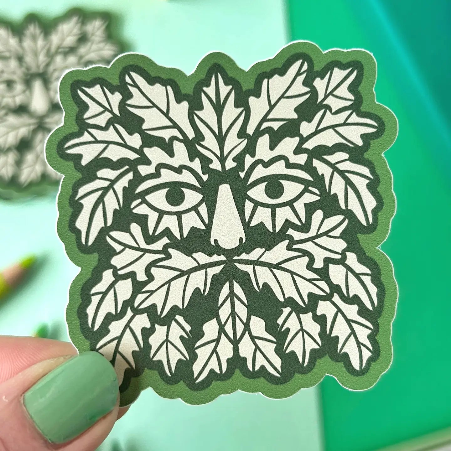 Hand displaying Stacey McEvoy Caunt Folklore Green Man Sticker with oak leaf face on vibrant green background, eco folklore art.
