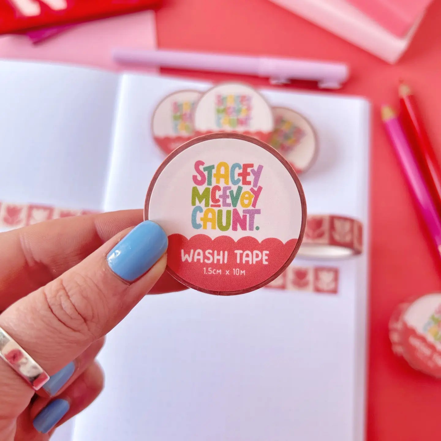 Hand displays Stacey McEvoy Caunt Red Folk Tulips washi tape above an open notebook with colorful pens on a workspace table.