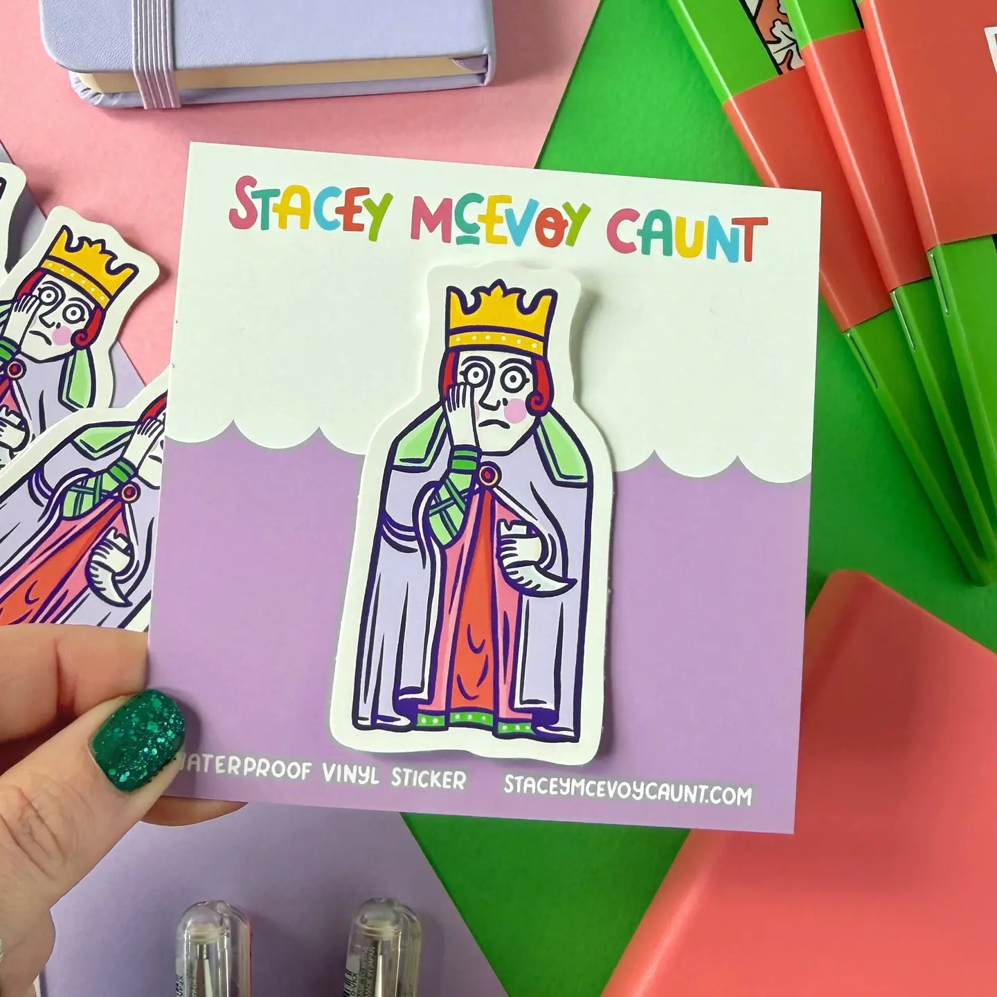 Hand holding Stacey McEvoy Caunt Medieval Queen Lewis Chessmen sticker, with colorful stationery items in the background.