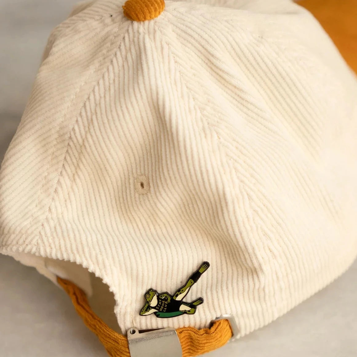 Forager Provisions LTD Stay Home Club enamel pin featuring a green frog exercising, attached to a cream corduroy cap.