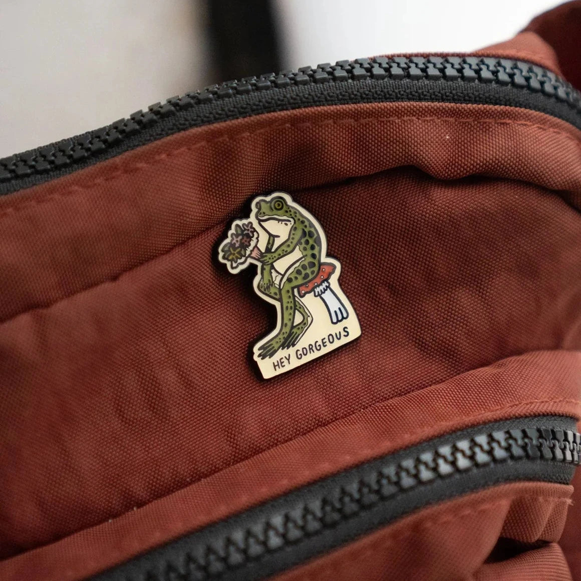 Stay Home Club Hey Gorgeous Pin decorates a brown bag near the zipper—chic positive accessory, uplifting motivational enamel pin.