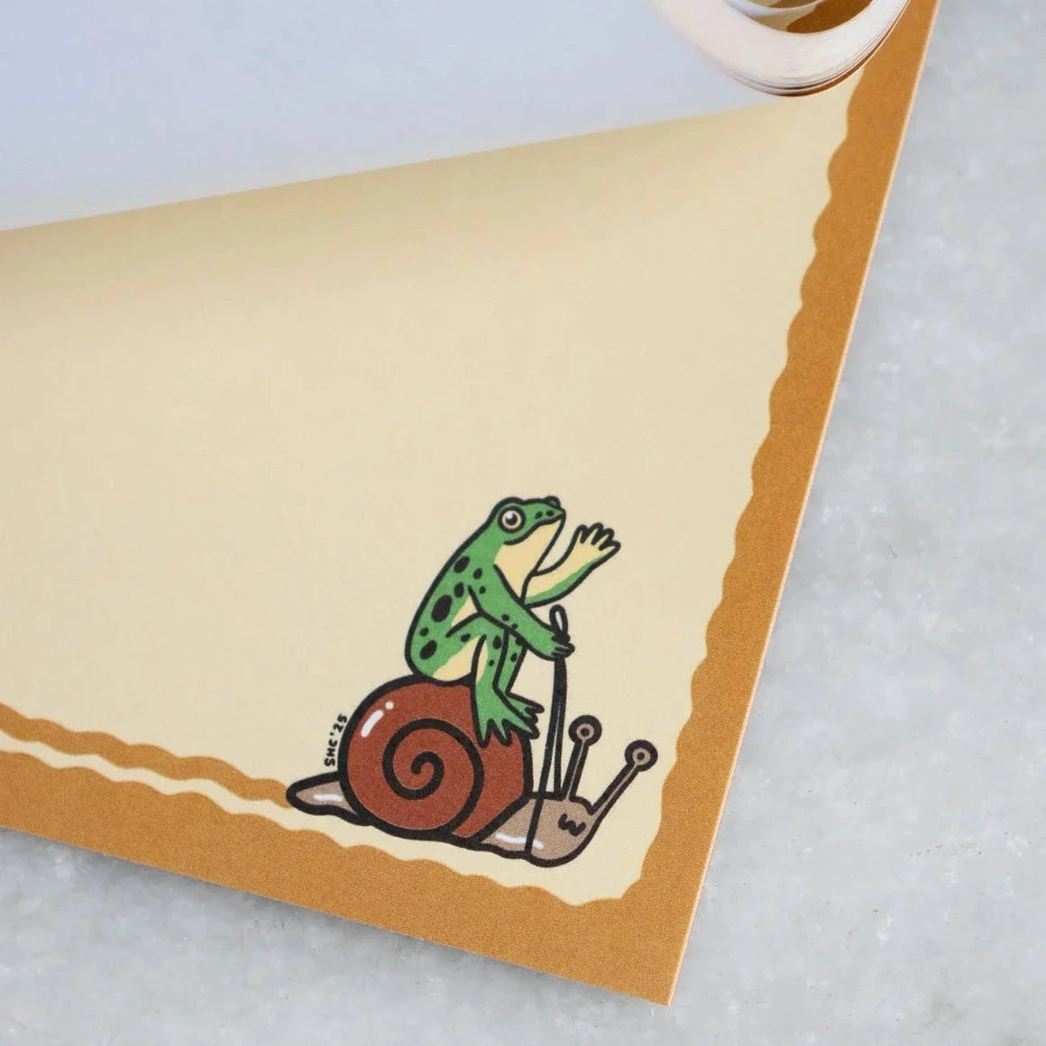 Green frog on brown snail illustration in the corner of Stay Home Club Frog & Snail Ride Notepad, cute stationery design.