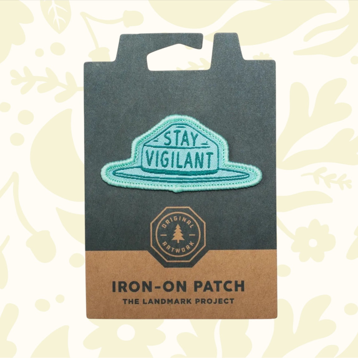 Teal iron-on patch featuring the phrase "STAY VIGILANT." Ideal for outdoor adventures, the packaging highlights "The Landmark Project," "Original Artwork," and includes sustainable materials. Official product name: The Landmark Project Smokey Bear Patch - Smokey Hat.