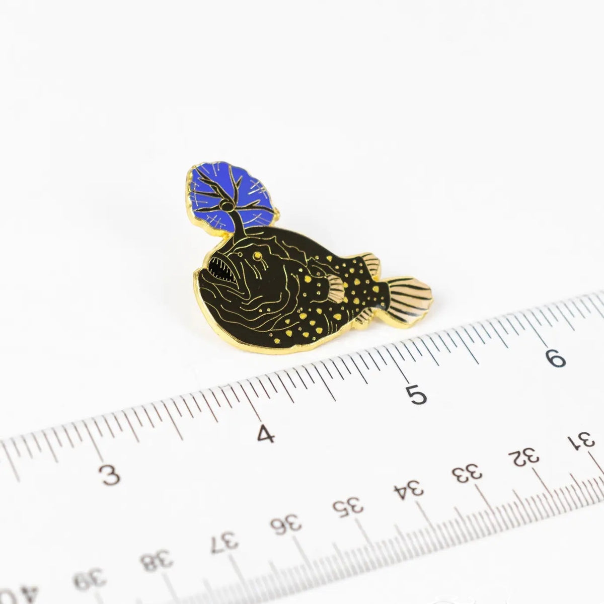 Stemcell Atlantic Football Fish enamel pin with a blue lure next to a ruler on white background, collectible jewelry accessory.