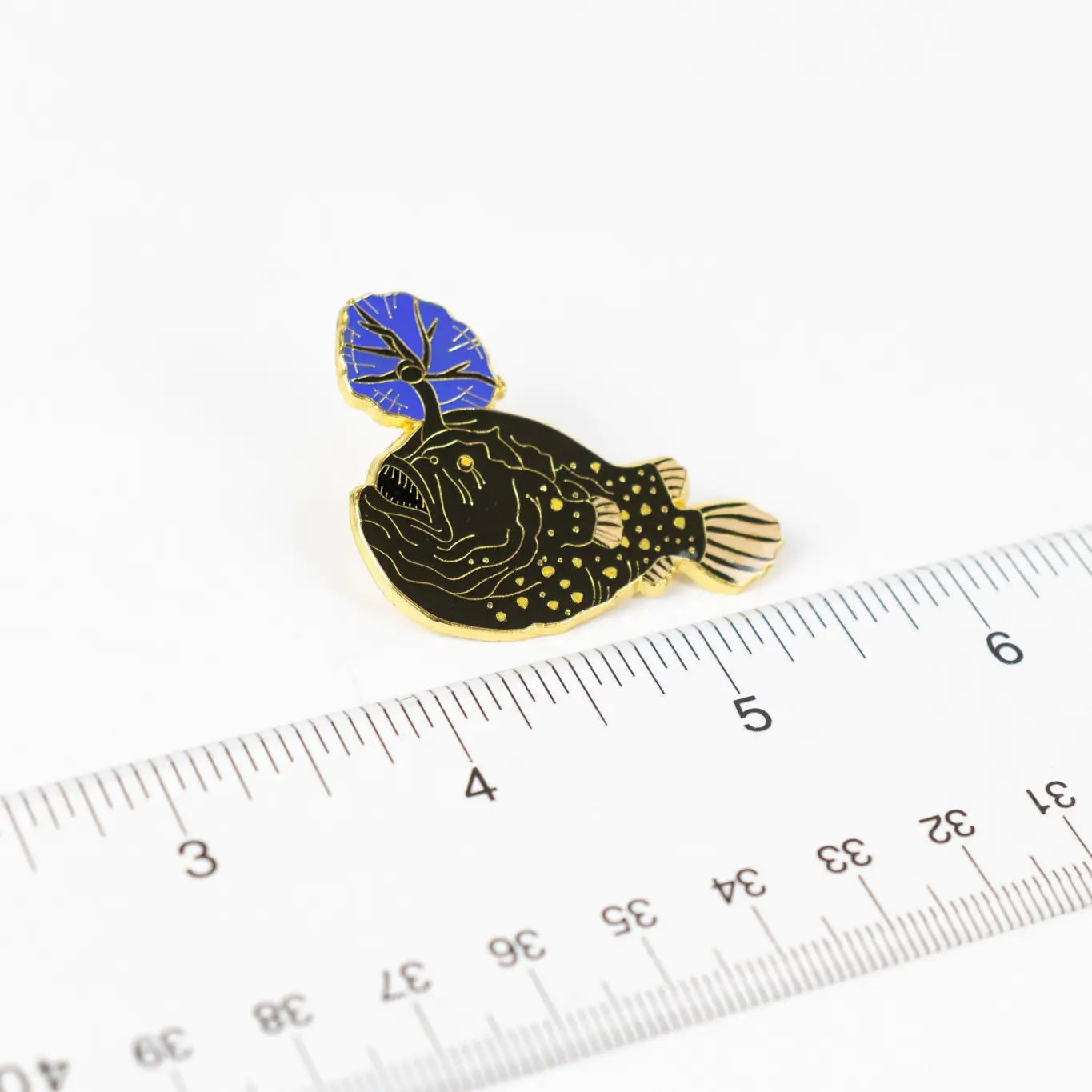 Stemcell Atlantic Football Fish enamel pin with a blue lure next to a ruler on white background, collectible jewelry accessory.