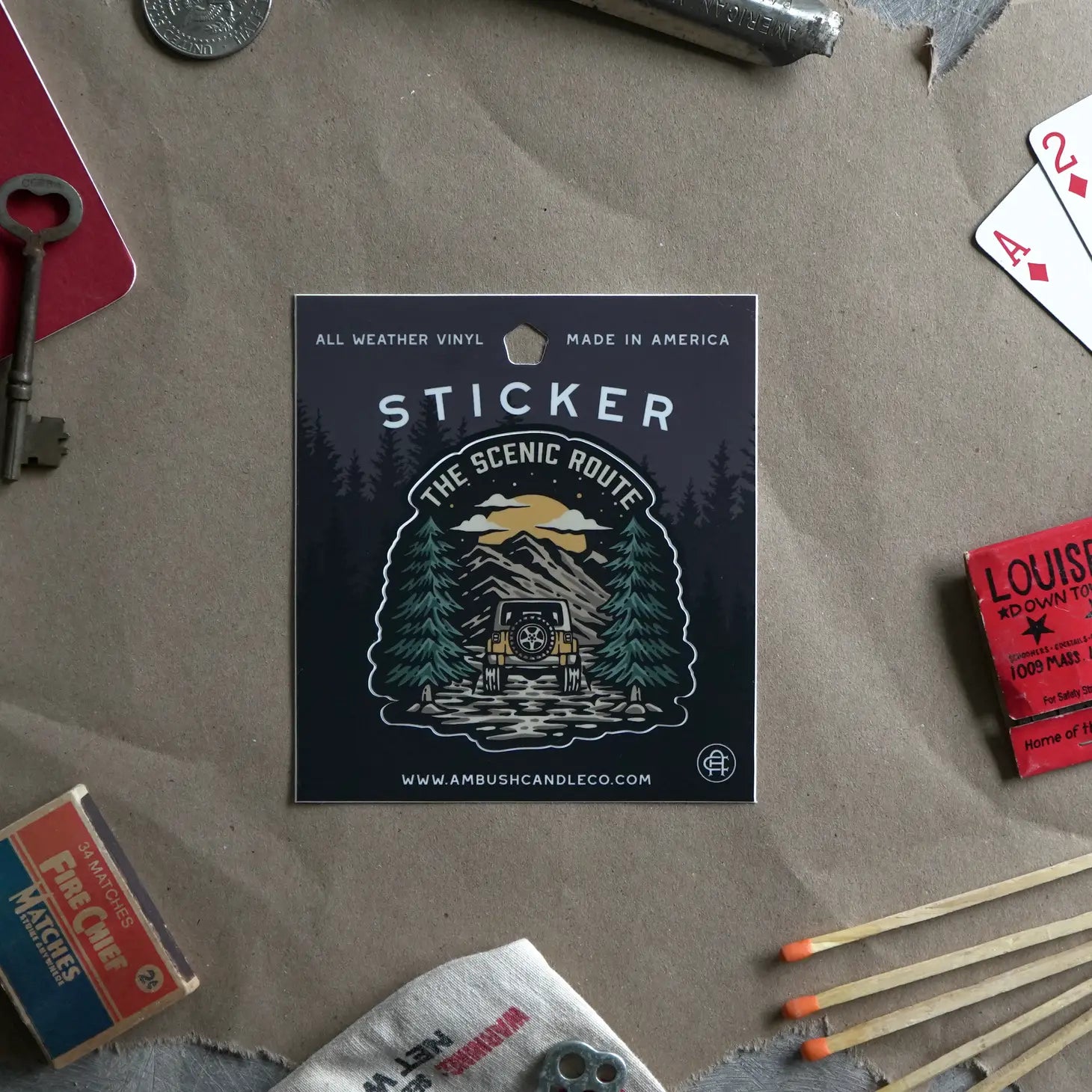 An Ambush Candle Co The Scenic Route Sticker, featuring a weatherproof vinyl design with a snowy cabin scene, is placed on ground covered in dried autumn leaves. Made in the USA, this high-quality sticker is also dishwasher safe.