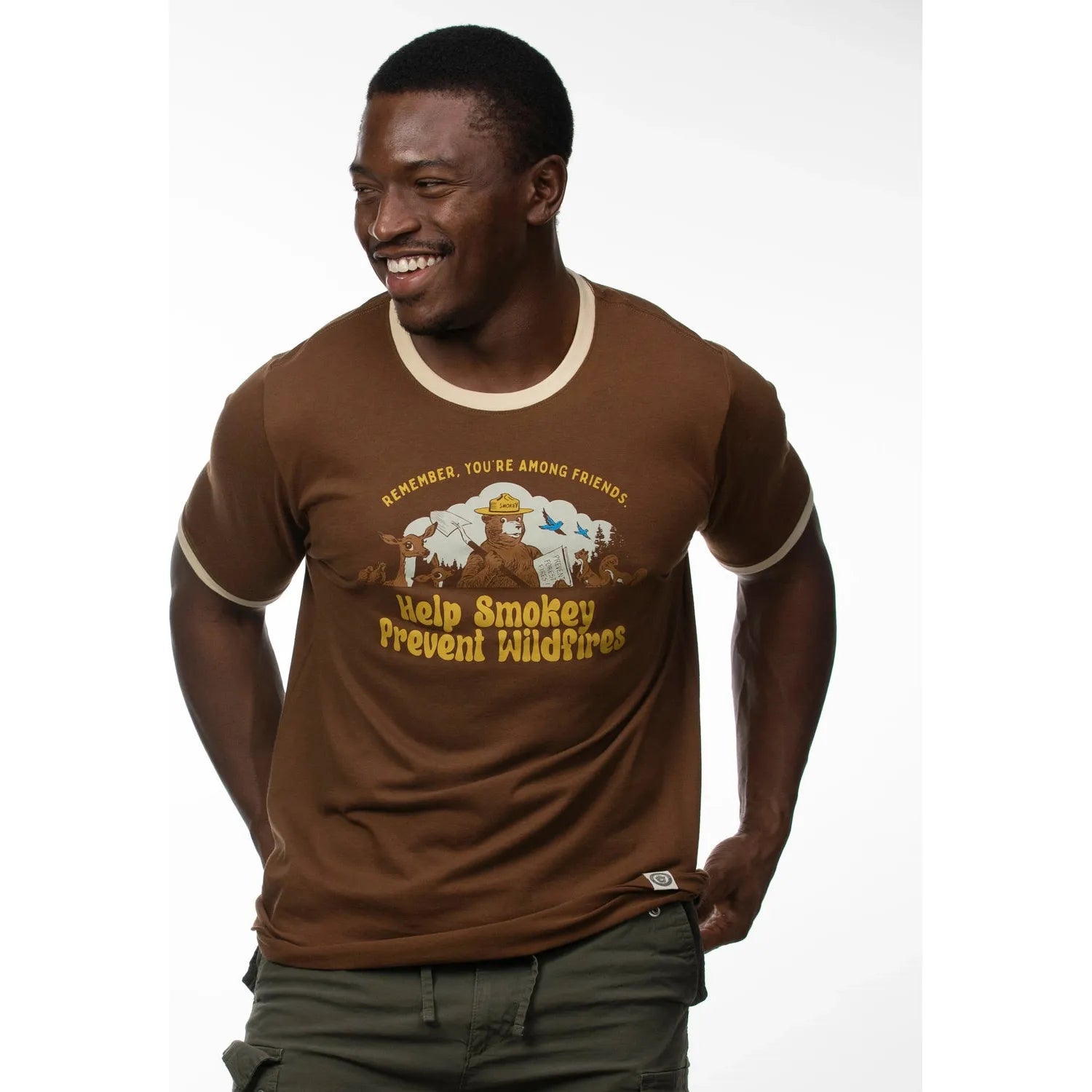 A smiling man wearing a walnut "The Landmark Project Amongst Friends Smokey Bear T-Shirt" with khaki pants against a plain white background, showcasing Smokey Bear products that support wildfire prevention education.