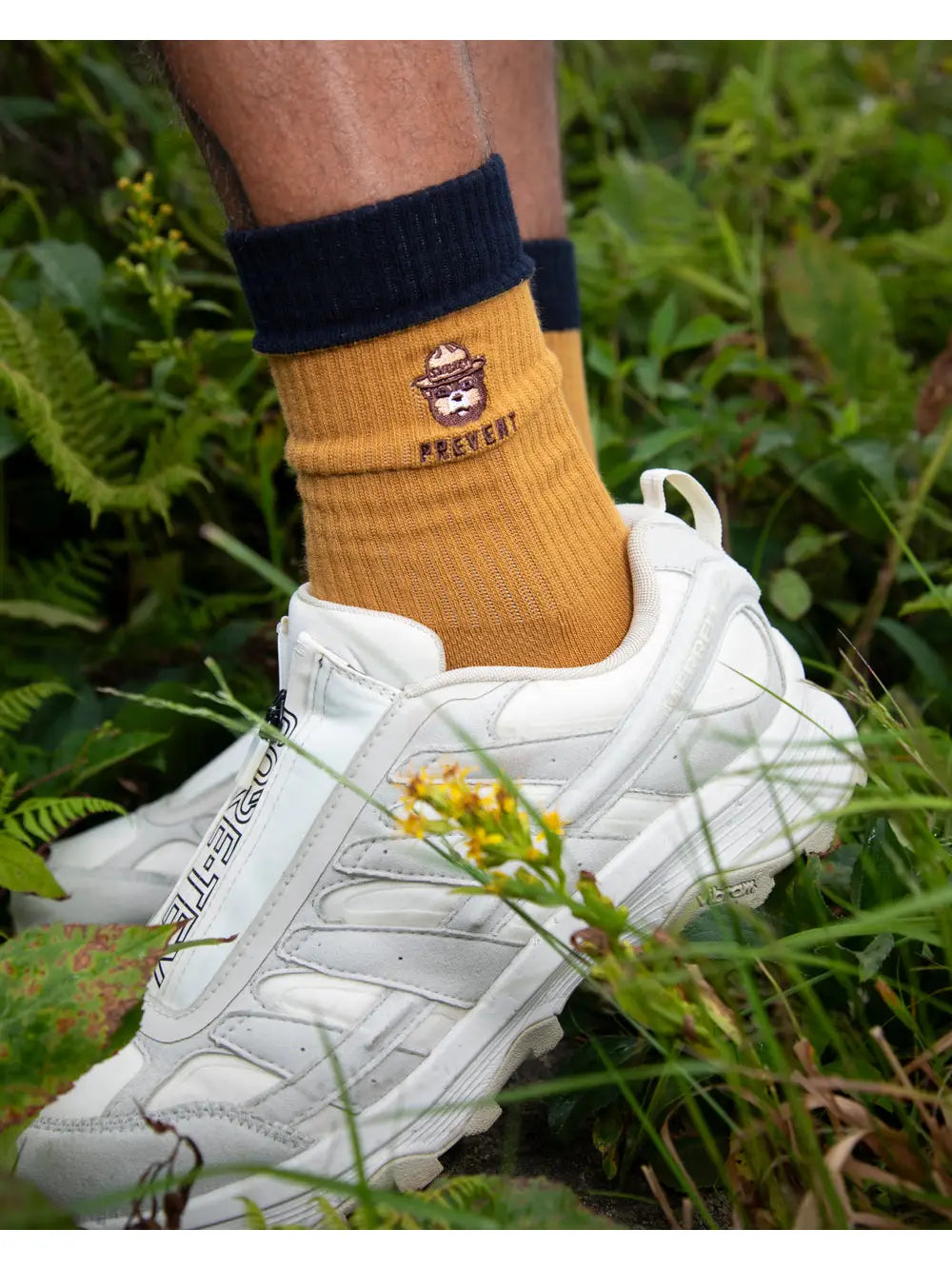 Person wearing white sneakers and Landmark Project Smokey Bear "PREVENT" socks standing on lush green grass outdoors.