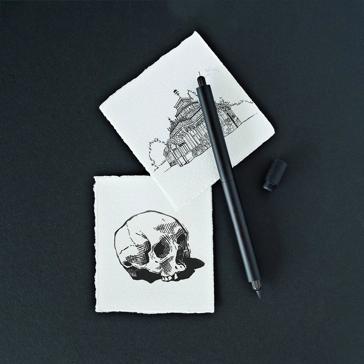 Two ink sketches—a human skull and an old building—next to Tom’s Studio Lumos Pro Duo Black Pen on black desk.