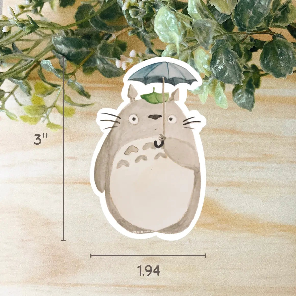 An adorable Totoro vinyl sticker, featuring a cartoon-like, chubby grey creature with a white belly holding a tiny umbrella and surrounded by green leaves, designed for fans of Studio Ghibli. Get your Seenee Scribbles Studio Ghibli Inspired Totoro Sticker today!