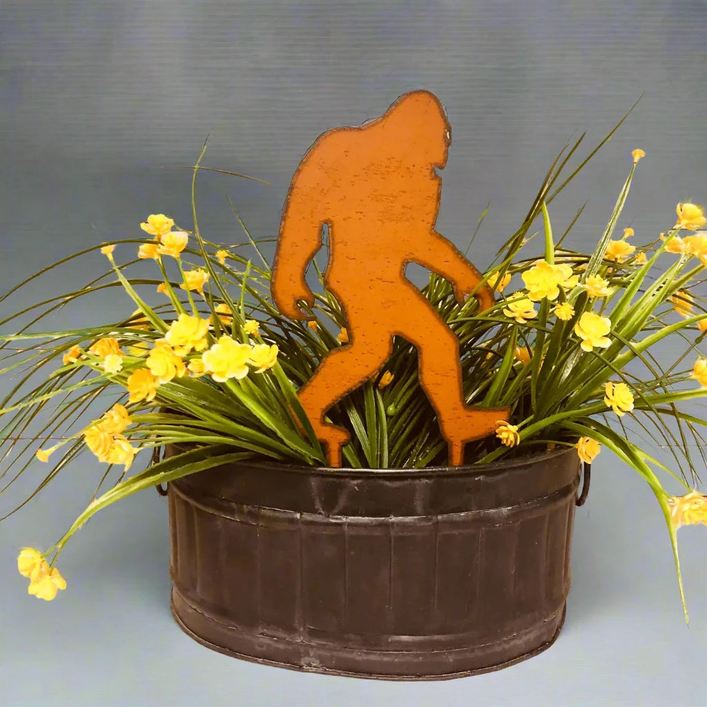 A rust-colored Universal Iron Works Bigfoot Yeti Walking Stake Planter Decoration stands among green grass and yellow flowers in a black metal planter against a wooden background, making it a charming piece of garden decor.