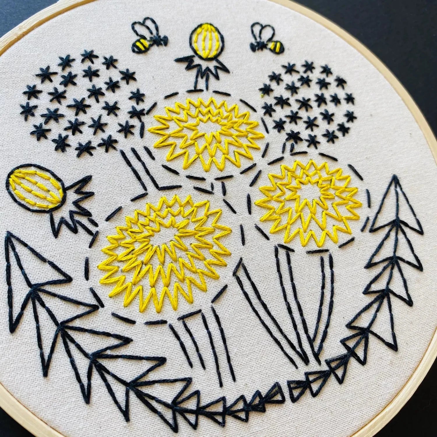 The Hook Line & Tinker Complete Embroidery Kit - Dandelion showcases beige fabric adorned with yellow flowers, black stems, leaves, and two bees. This beginner-friendly embroidery kit is entirely plastic-free.