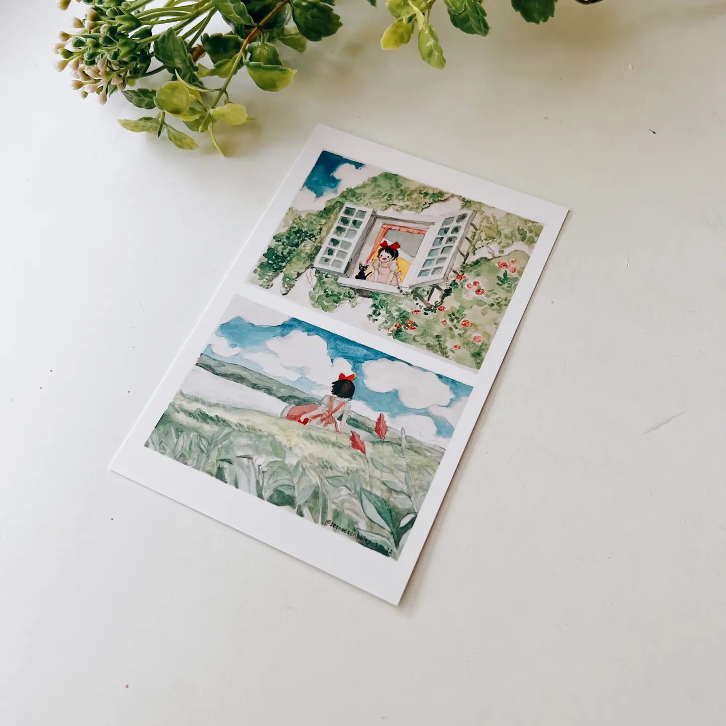 A hand holds the Seenee Scribbles Art Print 4 x 6 - Kiki On A Clear Day, featuring two beautiful watercolor illustrations: a girl at a flowered window (top) and a girl in a field with birds (bottom), evoking scenes from Kiki's Delivery Service.